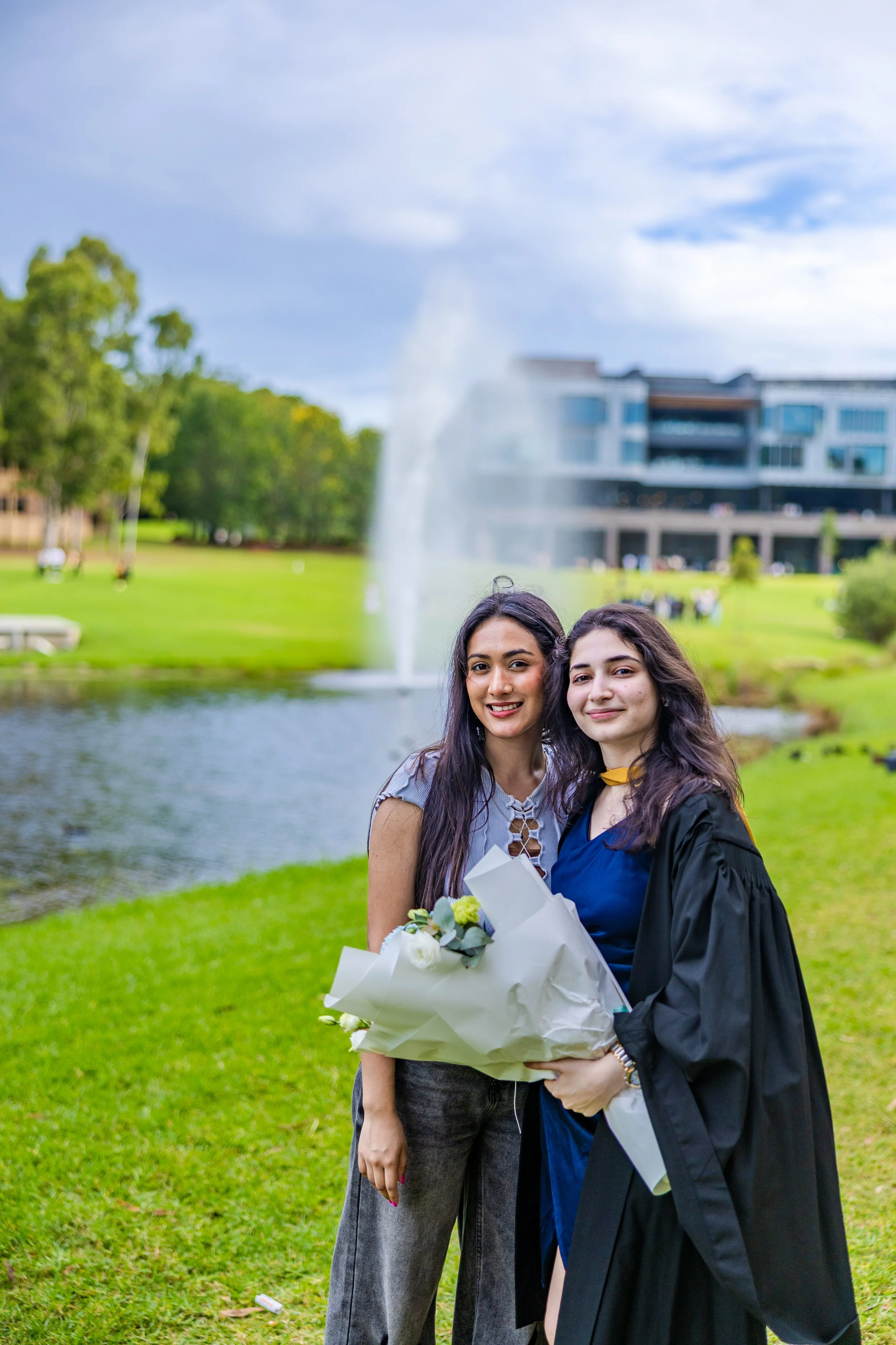 Two women standing together outdoors near a pond with a fountain, one in a graduation gown holding a bouquet of flowers, the other smiling beside her.