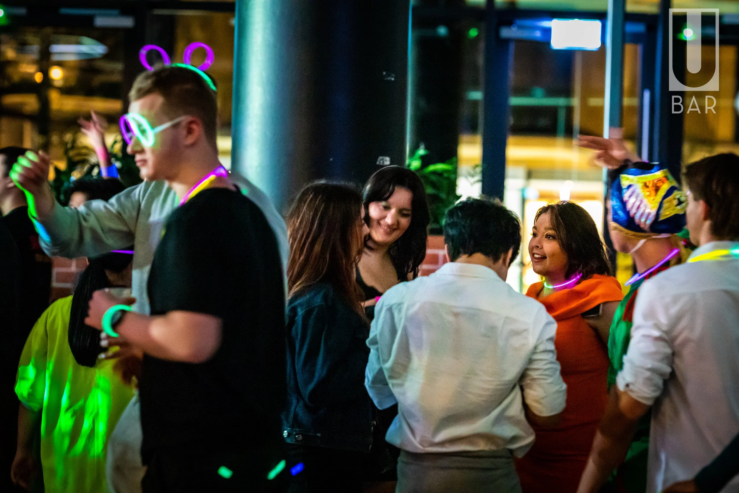 Group of people dancing and socializing at a neon-themed party, some wearing glow-in-the-dark accessories.