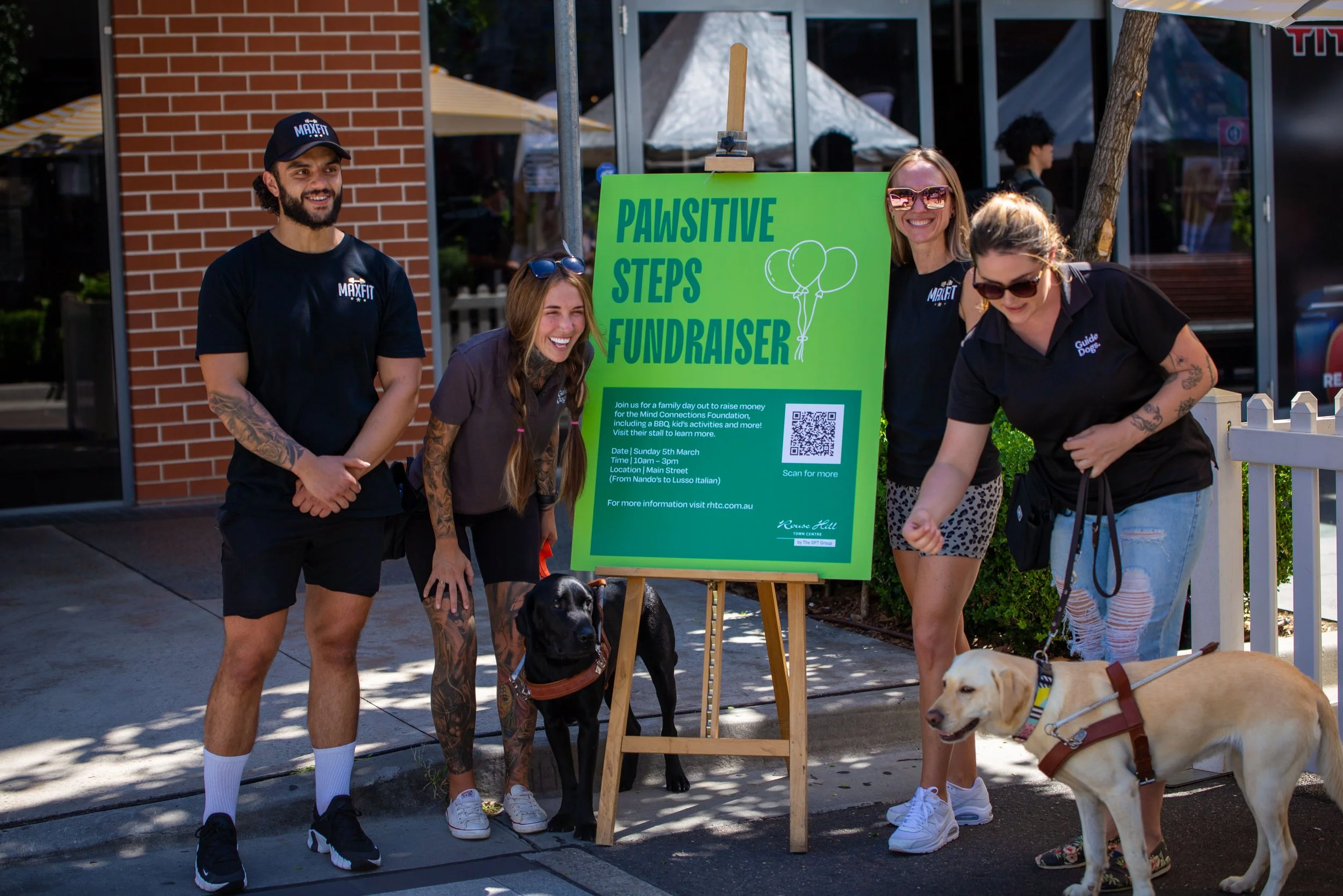 Group of four people, two women and two men, standing outdoors next to a bright green sign that reads 'Pawsitive Steps Fundraiser,' with two dogs. The people are smiling, and it's a sunny day outside a brick building with tents in the background.