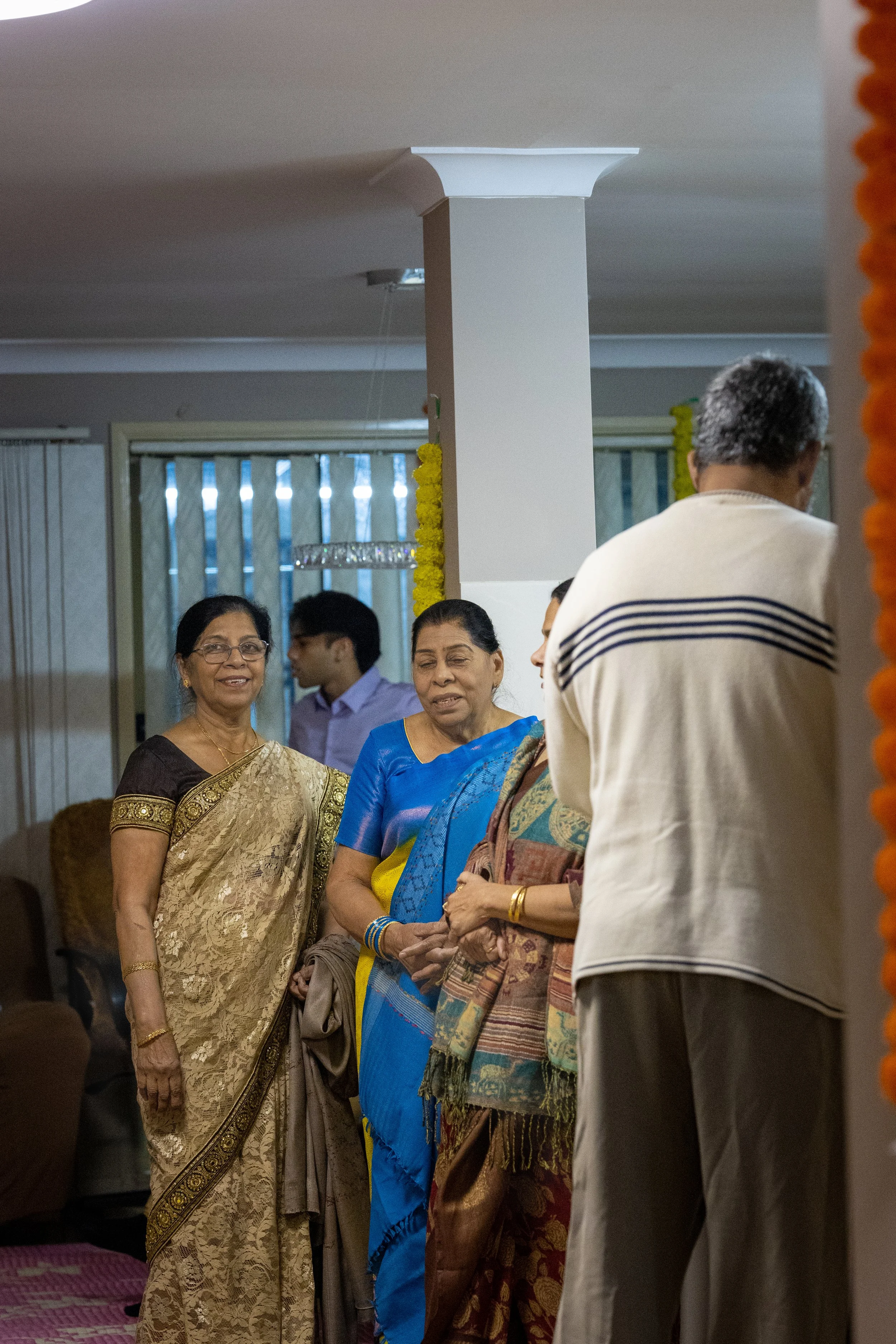 A group of Indian people, including women dressed in sarees and a man in a shirt, gathered in a living room, likely attending a family gathering or celebration.