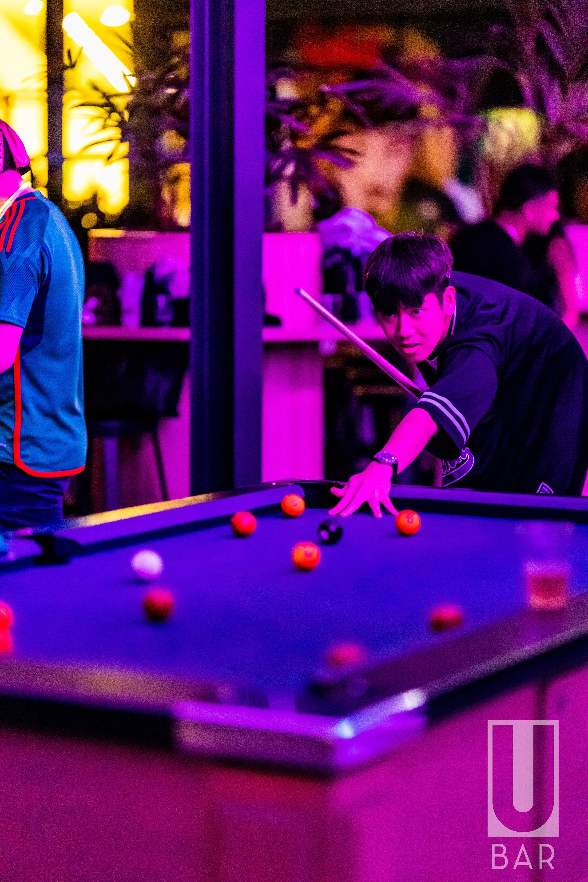 A young man playing pool at a bar with purple lighting, focusing on making a shot, with other patrons in the background.