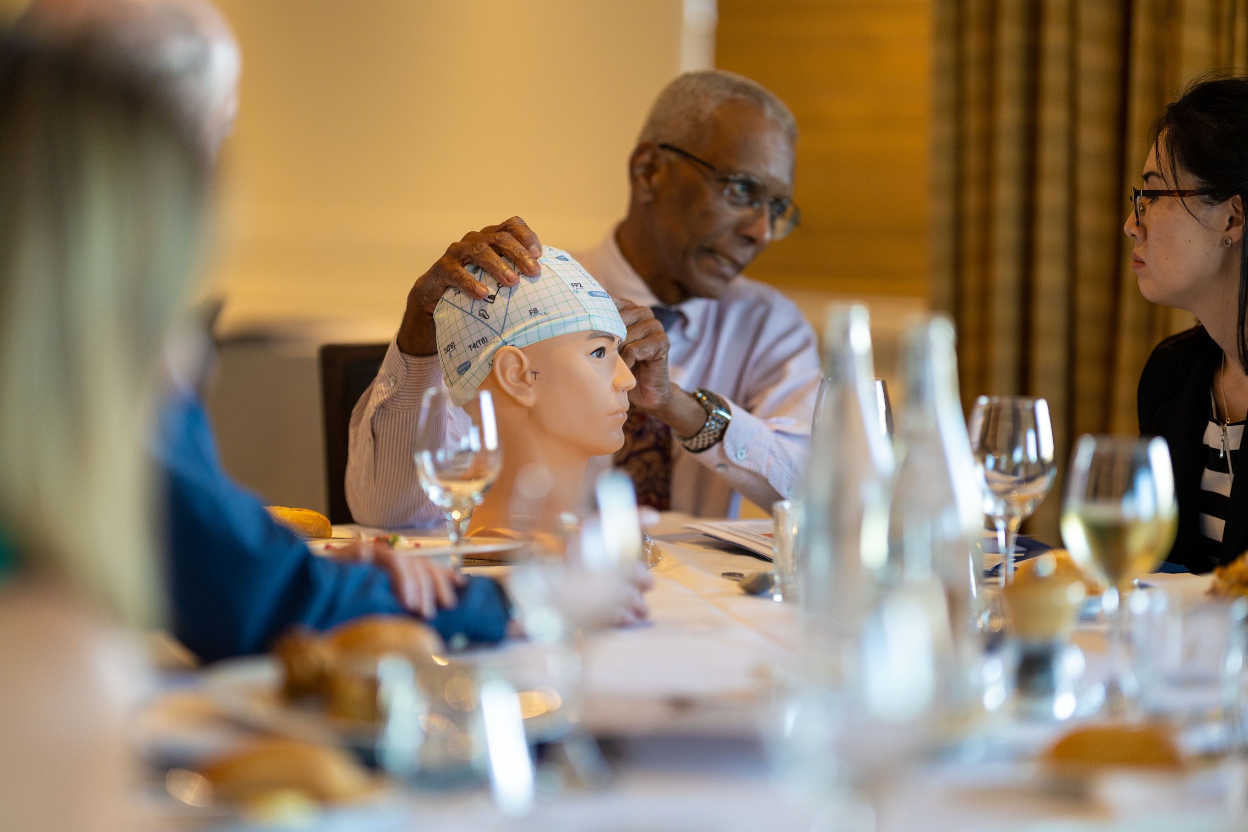 An elderly man wearing glasses is adjusting a medical mannequin's head, which is wearing a surgical cap, at a dining table set with glasses and plates, while other people look on during a demonstration or seminar.
