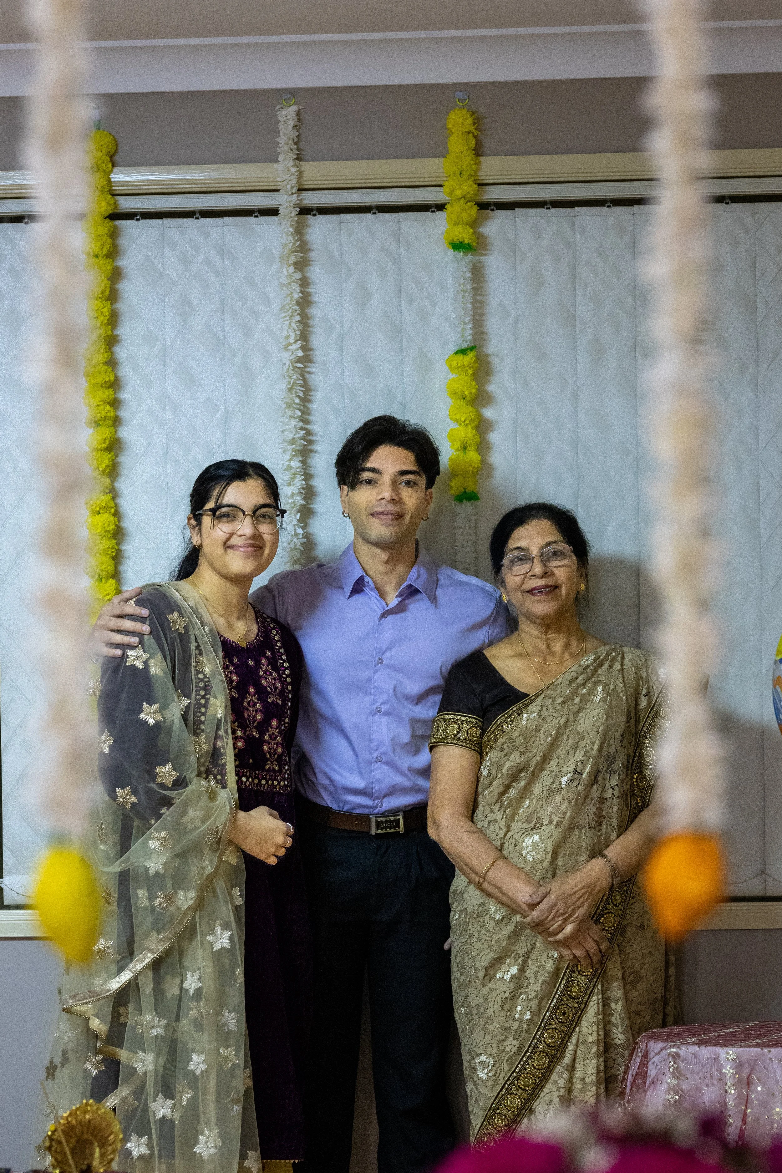 Three people posing in front of a decorated wall, with decorative hanging garlands behind them. An older woman in a gold and black saree, a younger woman in a traditional outfit, and a young man in a light purple dress shirt.