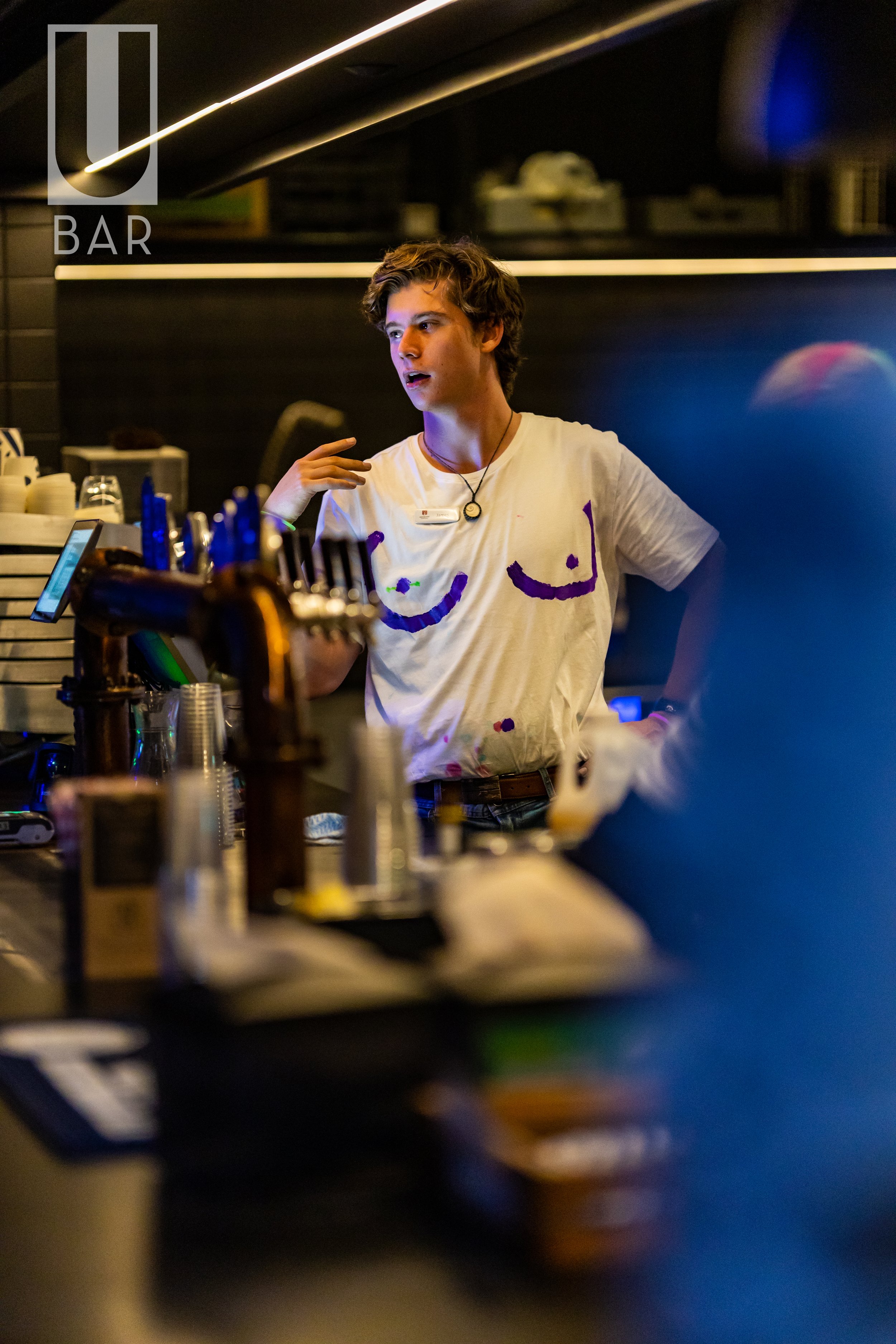 A young male bartender with curly hair and a necklace, wearing a white t-shirt with a face drawn on it, standing behind a bar counter with bottles and equipment, in a dimly lit bar with the sign 'U BAR' in the background.