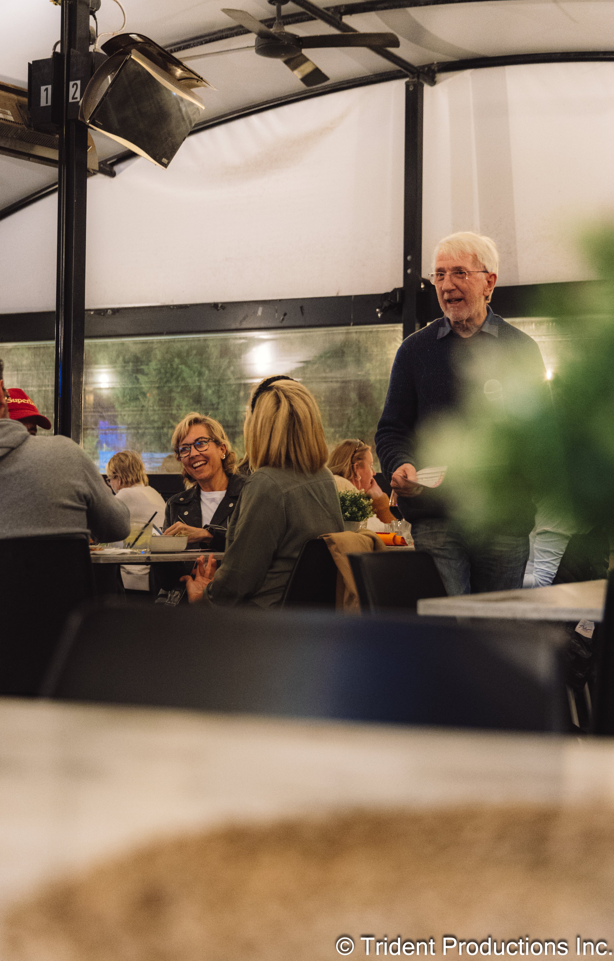 People sitting and talking at a restaurant or cafe, with a man standing and holding papers, smiling. The setting is indoors with a tent-like ceiling and large windows.