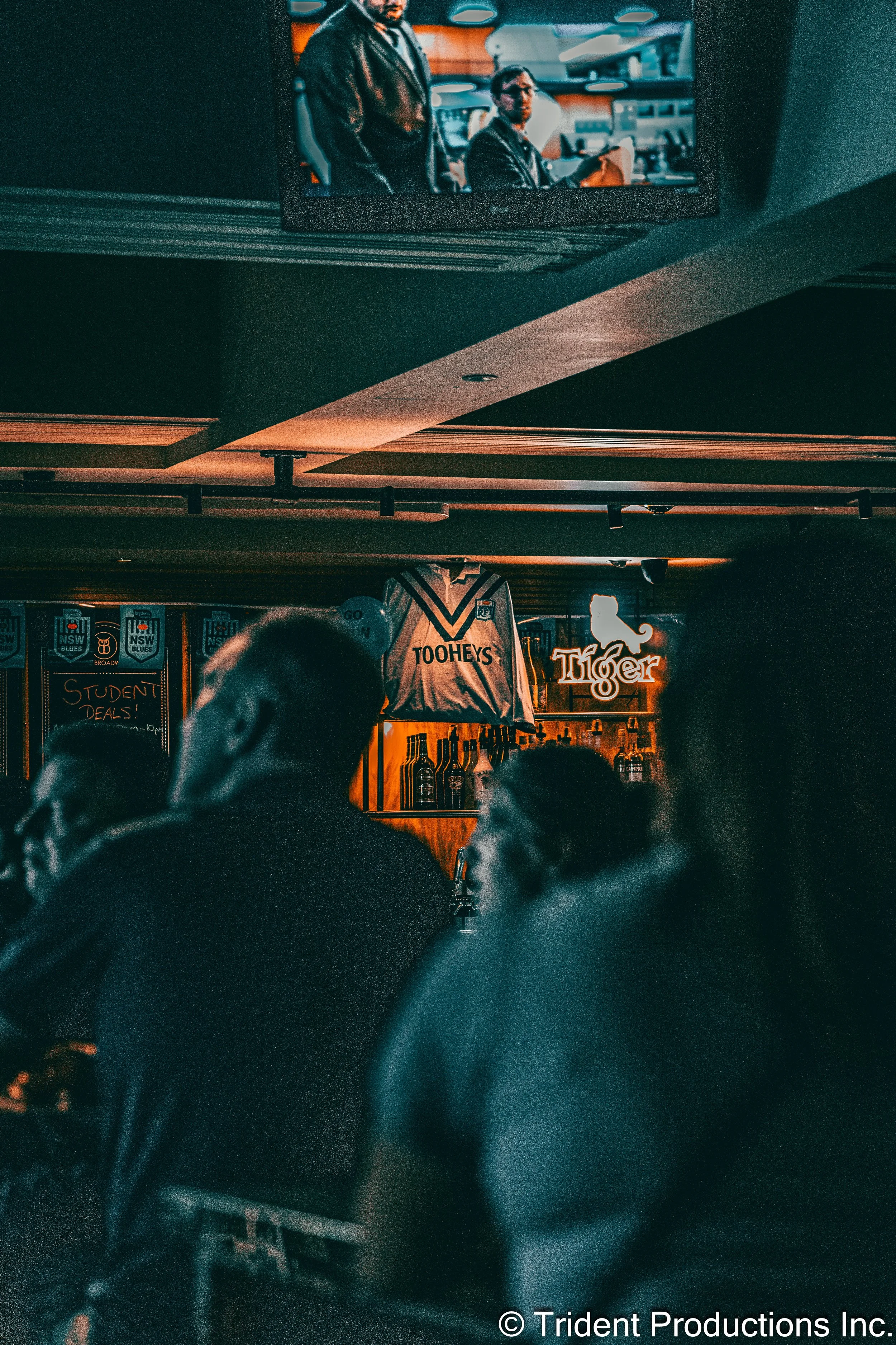 A dimly lit bar scene with people sitting at the counter, a jersey hanging on the wall with the word 'TOOHEYS,' and a neon sign shaped like the state of California with the word 'Tiger.' A television screen above shows two men, one standing and one s