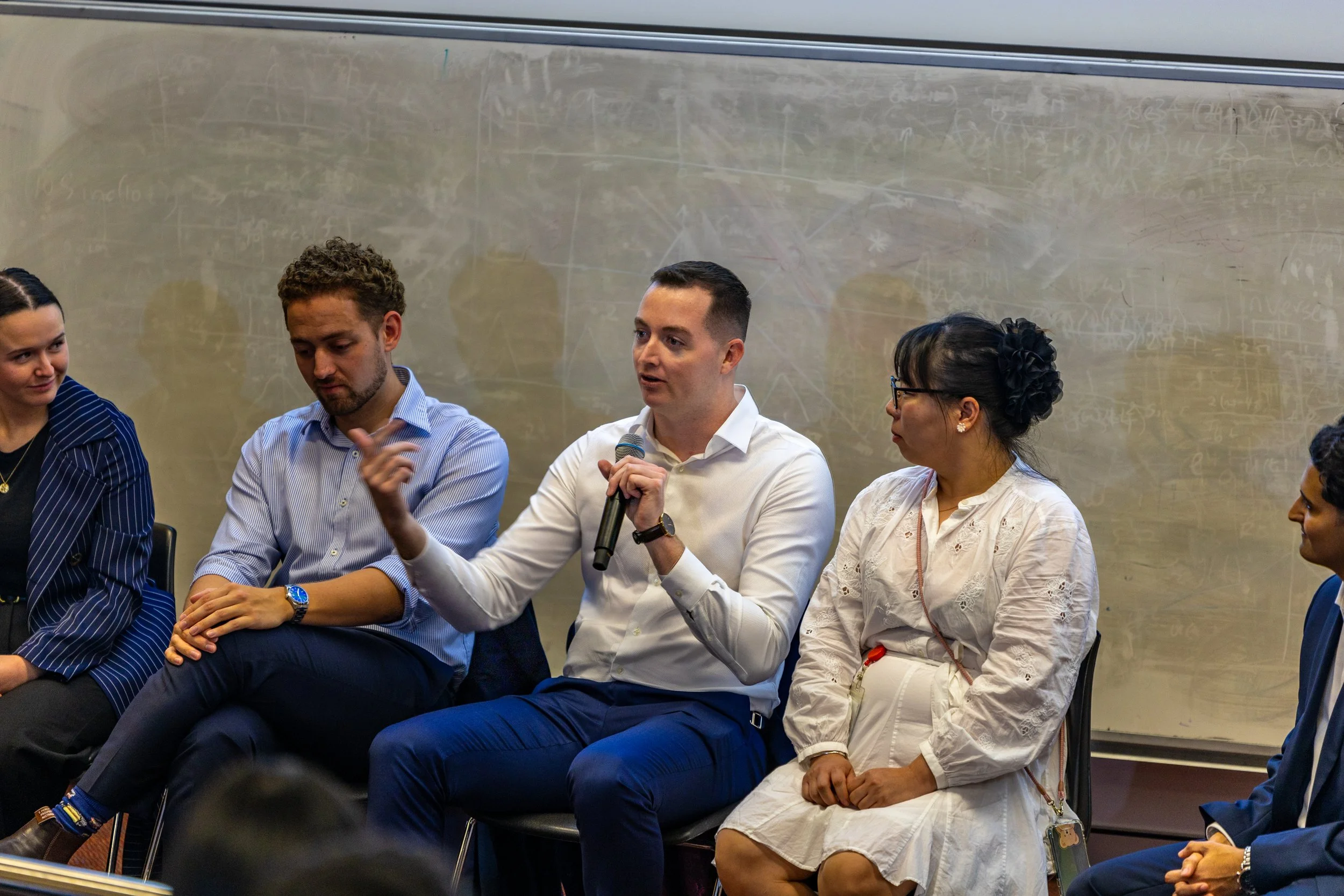 A group of five people sitting in a panel discussion, with a man in the center holding a microphone and speaking, in a room with a chalkboard in the background.