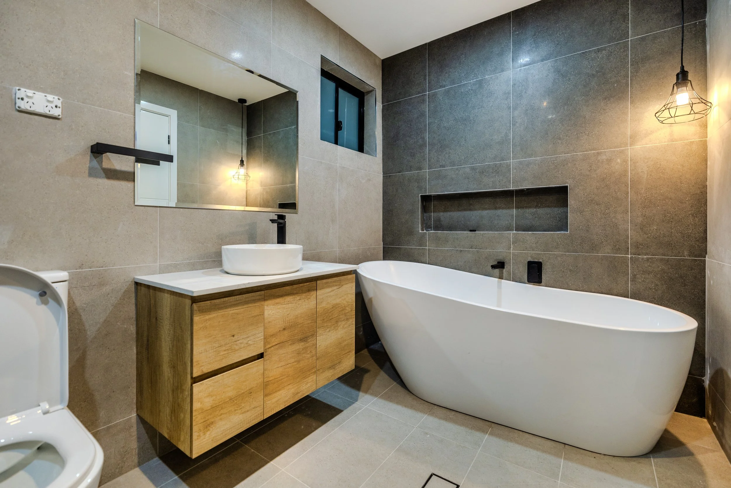 Modern bathroom with a white freestanding bathtub, a wooden vanity with a round vessel sink, a large mirror, grey tiled walls, a small window, and warm pendant lighting.