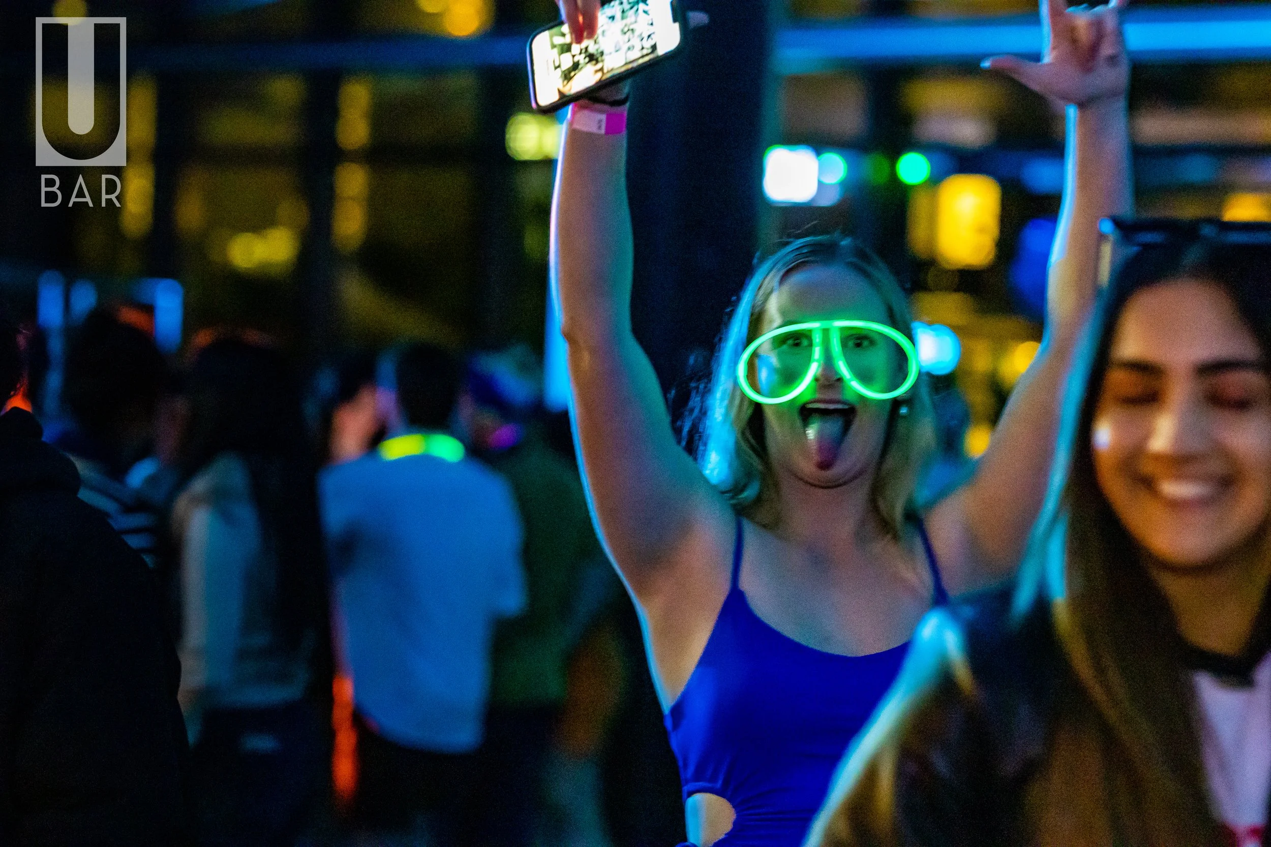 A woman at a lively party or club wearing neon green glasses and sticking out her tongue, with her arms raised, surrounded by people in a dark, colorful environment.