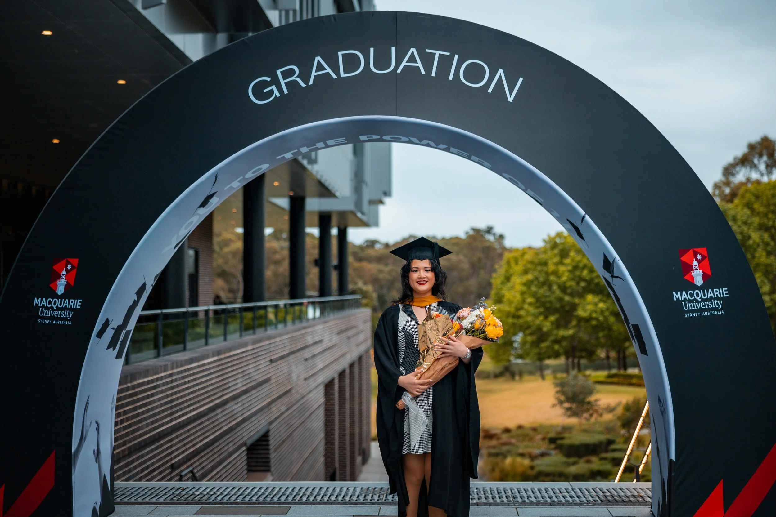 A woman in a graduation cap and gown holding a bouquet of flowers standing under a large Macquarie University arch that reads 'Graduation' in Sydney, Australia, with trees in the background.
