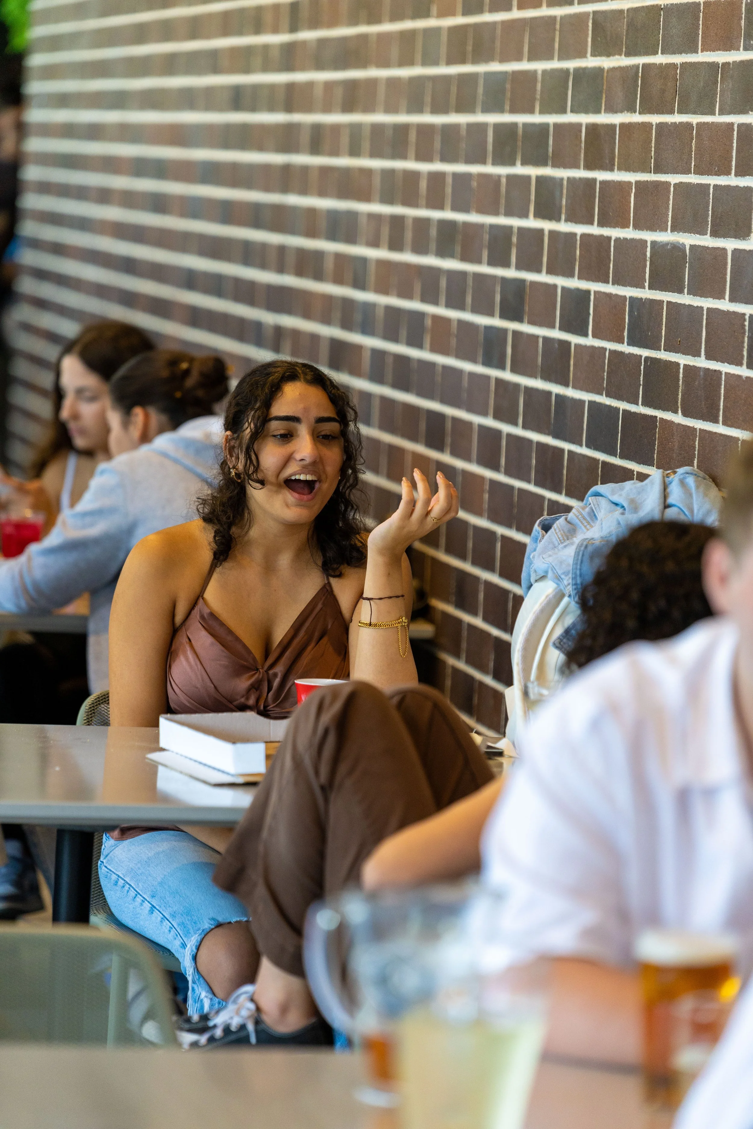 A young woman with dark, curly hair and a brown sleeveless top sitting at a table, talking animatedly with others nearby in a casual setting with a brick wall background.