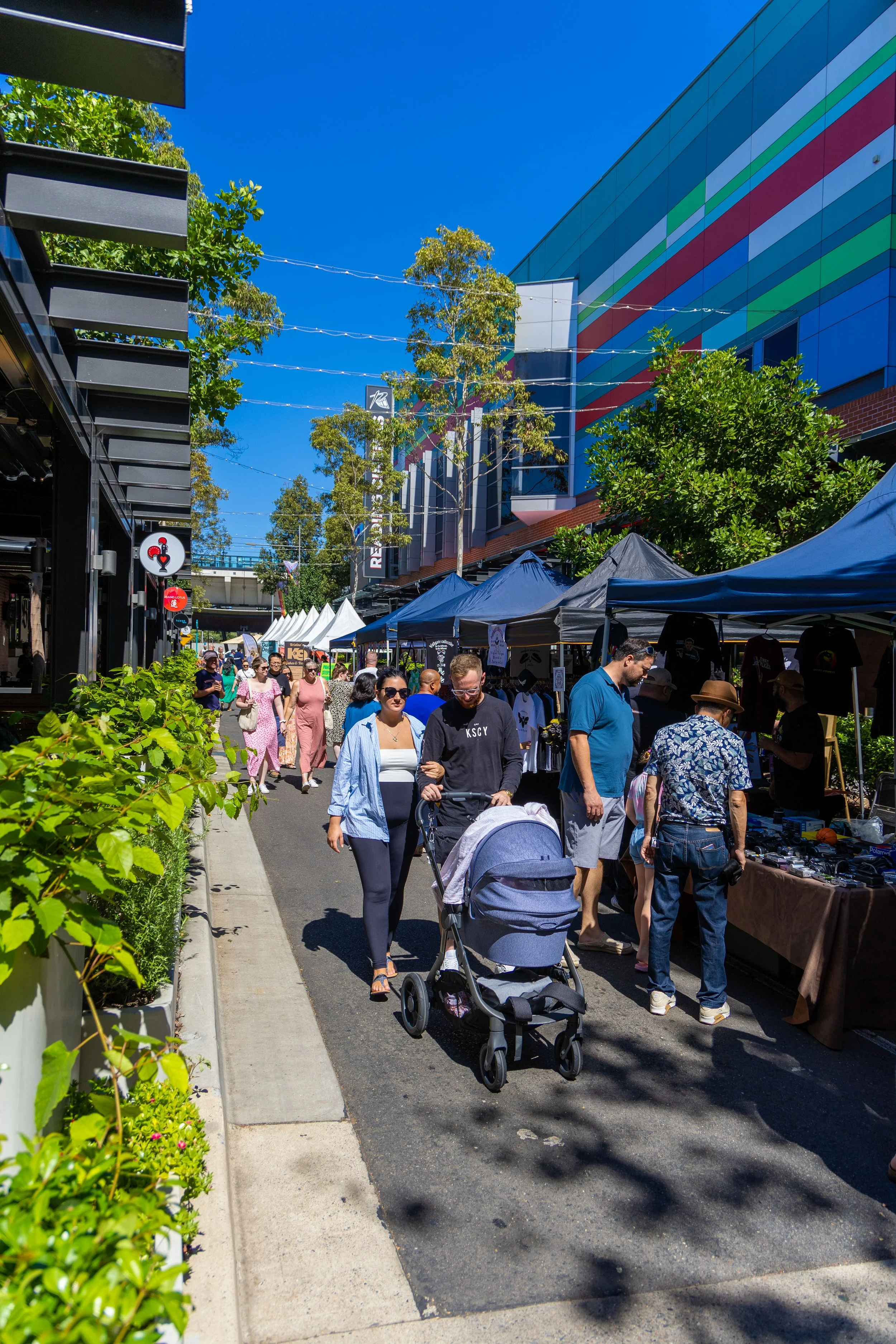People walking along a street market with tents selling items, trees, and a colorful building in the background on a sunny day.