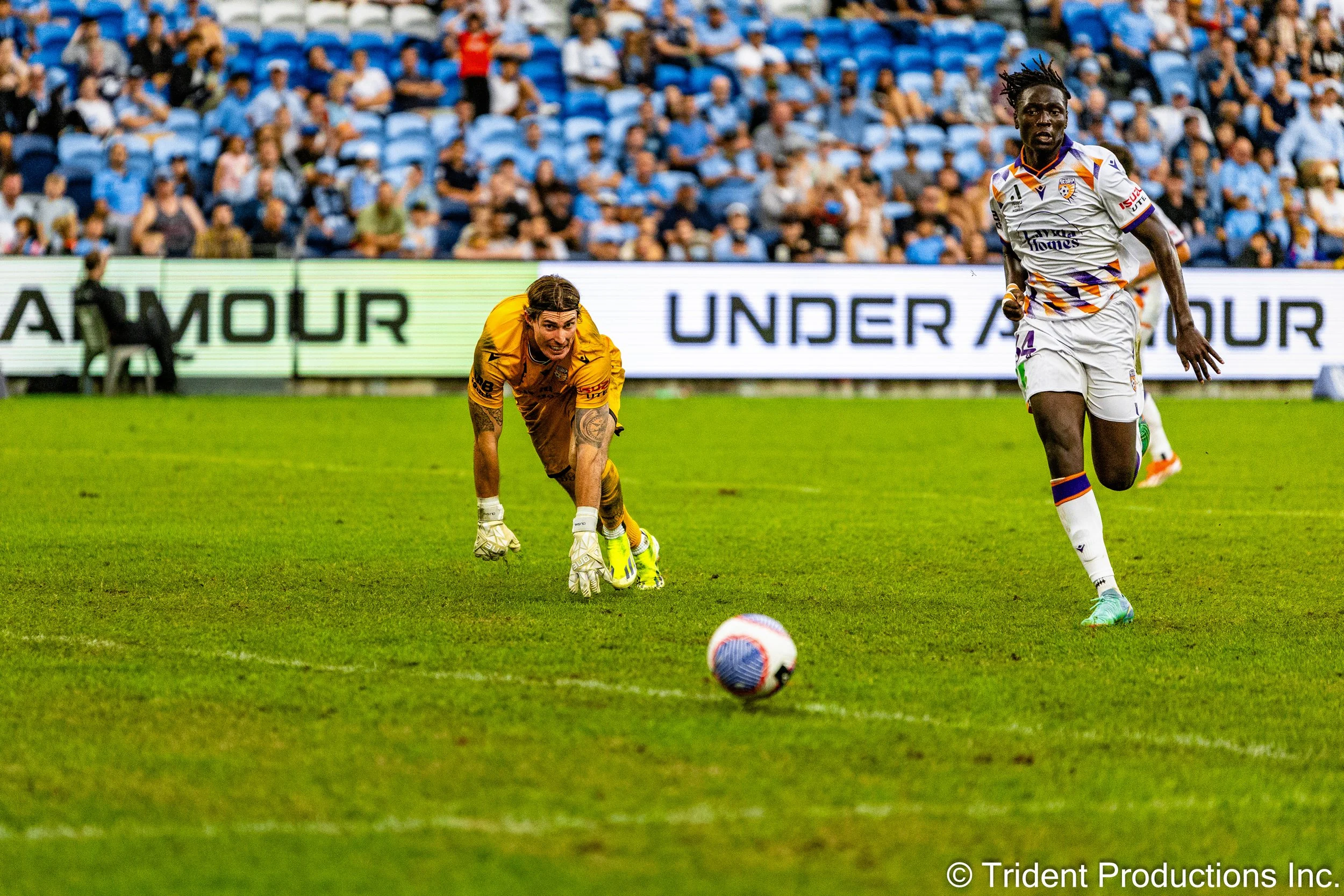 A soccer game with a goalkeeper in a yellow uniform on the ground and a player in a white uniform running after the ball on the field, with spectators in the stands in the background.