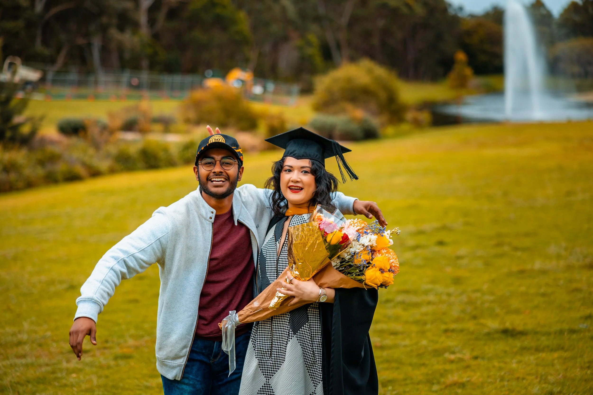 A woman in graduation attire holding a bouquet of flowers, standing with a smiling man outdoors in a park with a fountain in the background.