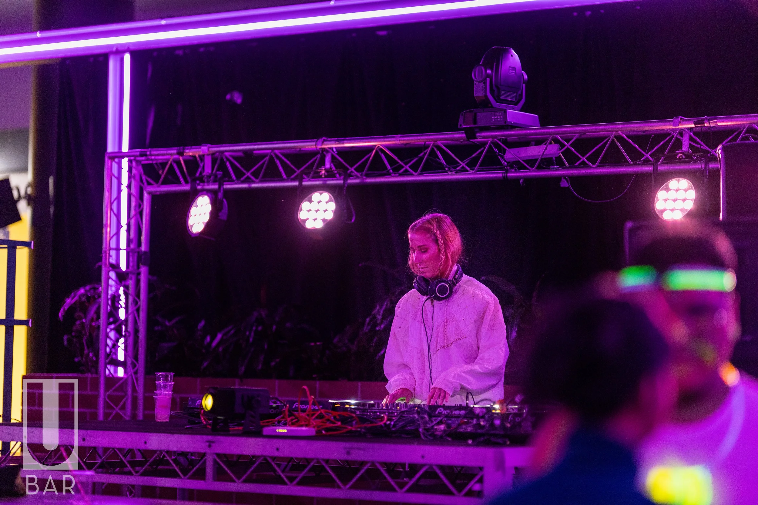 A female DJ wearing a white outfit and headphones, standing behind a DJ console with purple and pink stage lights illuminating her performance, in a nightclub or event setting.