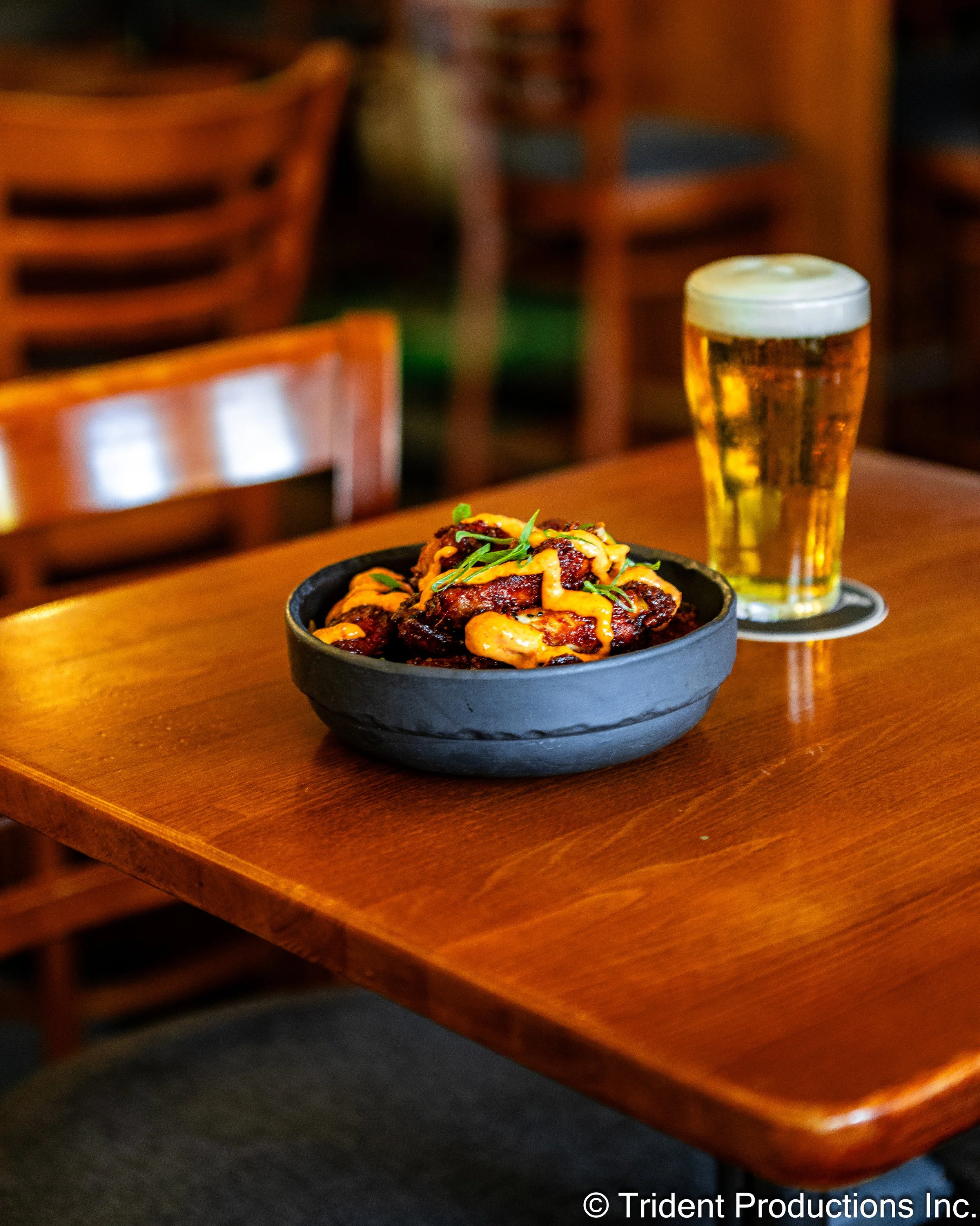 A bowl of spicy chicken wings topped with sauce and green garnish next to a glass of beer on a wooden table in a casual restaurant.