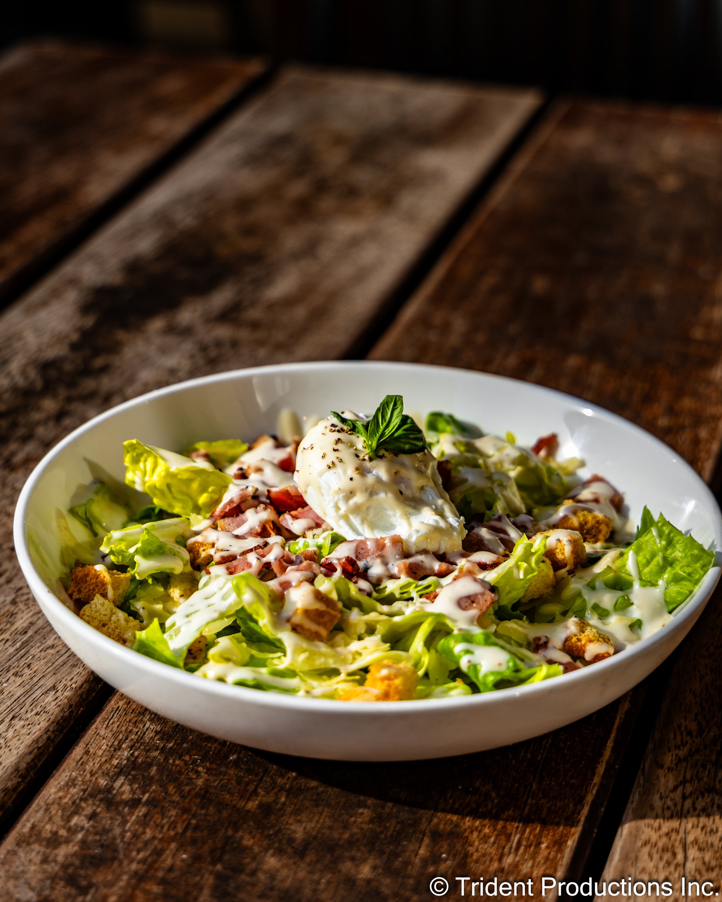 A Caesar salad with lettuce, croutons, bacon bits, a poached egg, and a basil garnish, served in a white bowl on a wooden table.