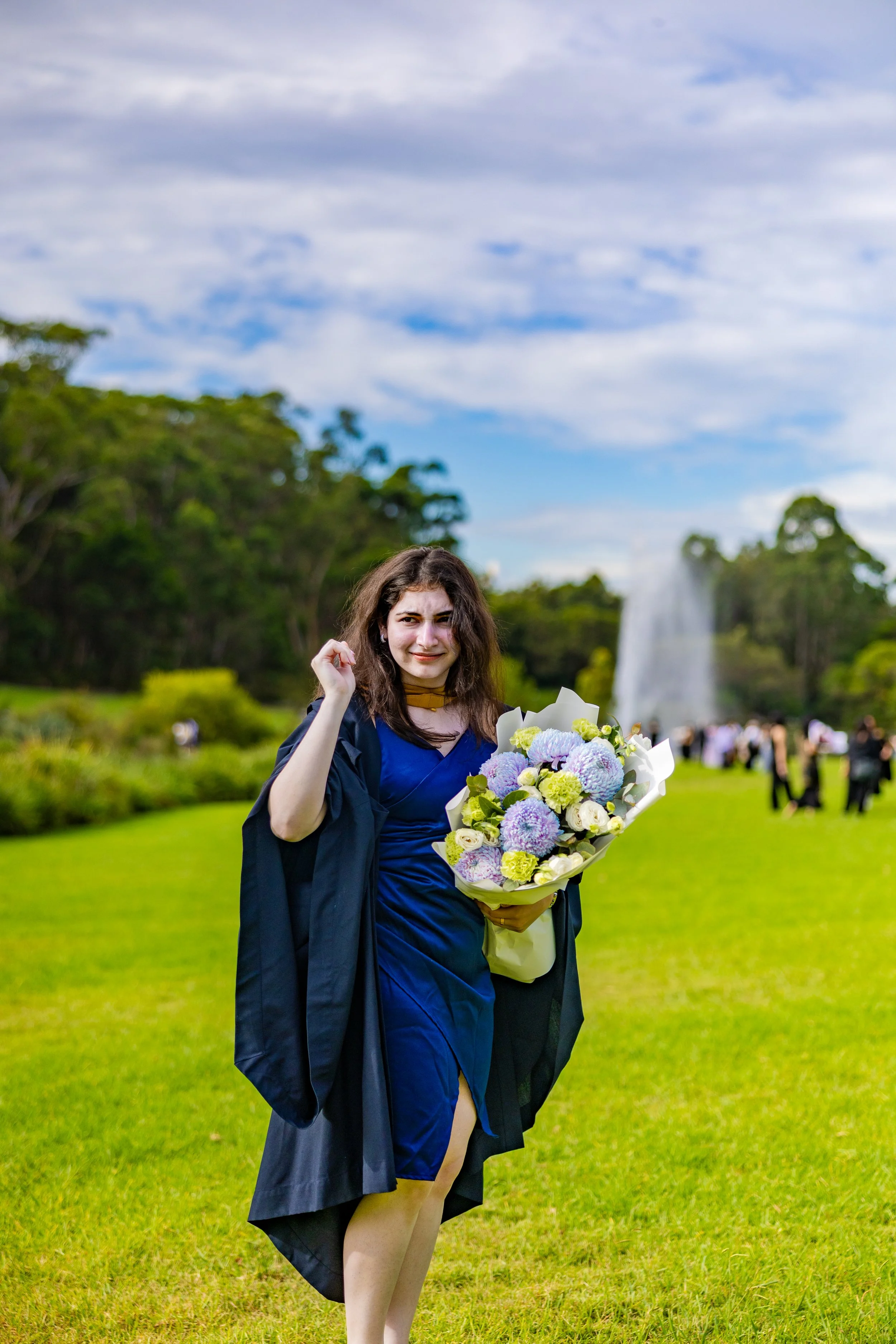 A young woman in a blue dress and a black graduation gown holding a bouquet of purple and white flowers, standing on a grassy area outdoors with other people and a large fountain in the background.