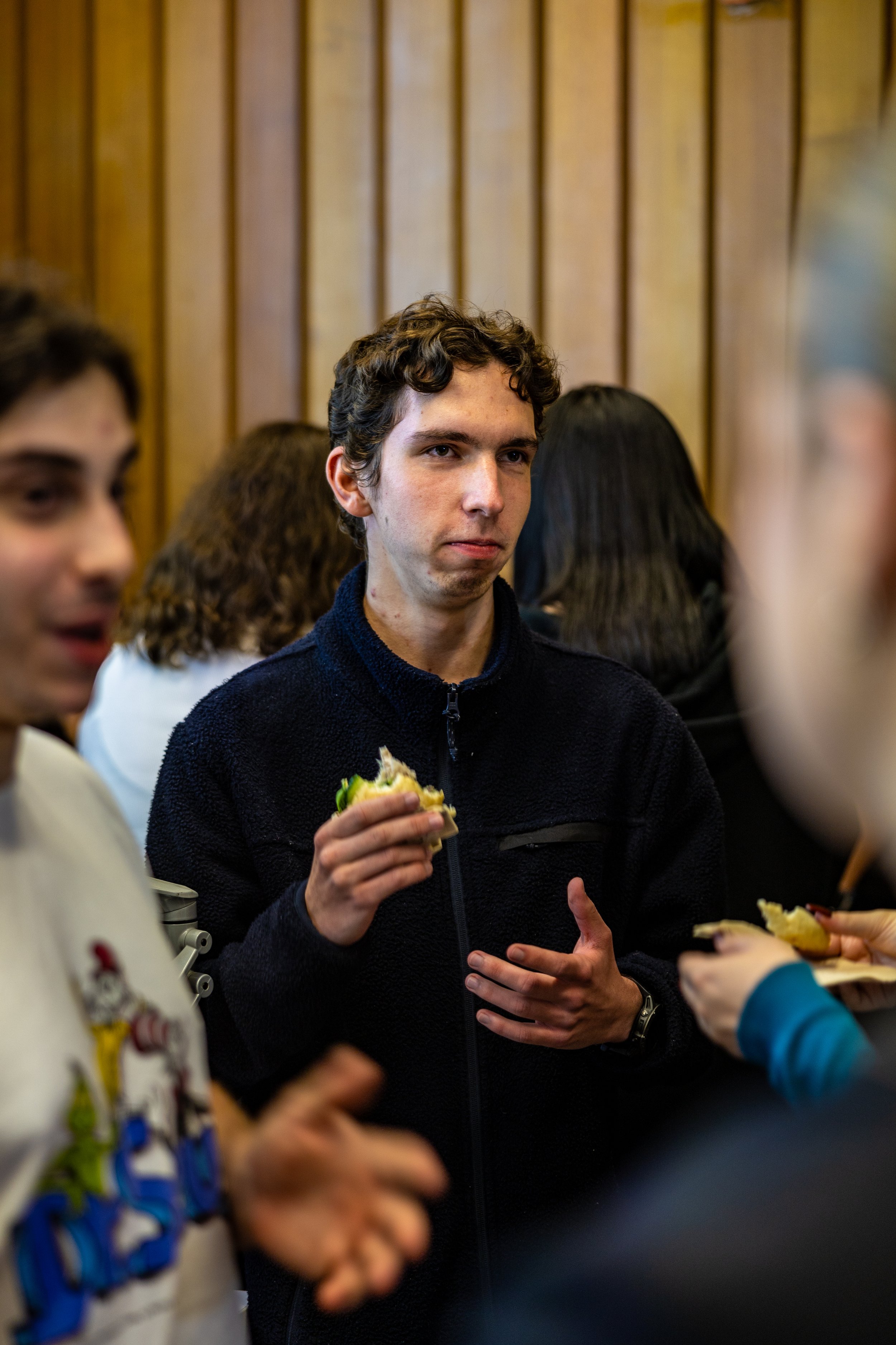 Man with curly brown hair in a black jacket holding a sandwich during a social gathering.