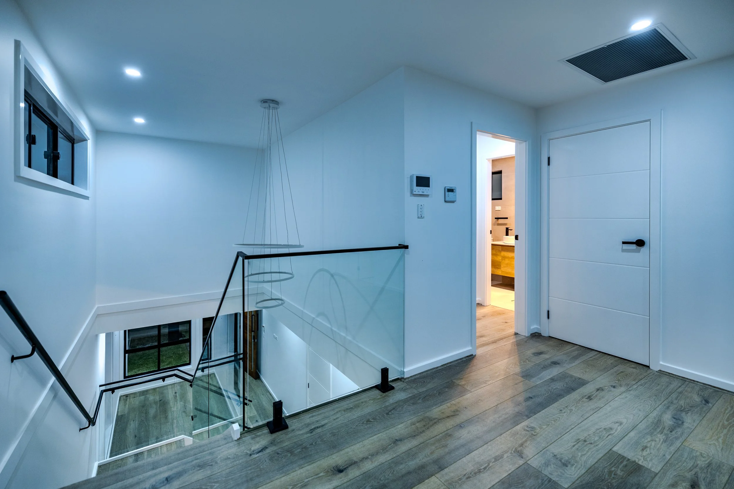 Interior view of a modern upstairs landing with white walls, wooden flooring, a staircase with glass and metal railing, small window, and an open doorway leading to a bathroom.