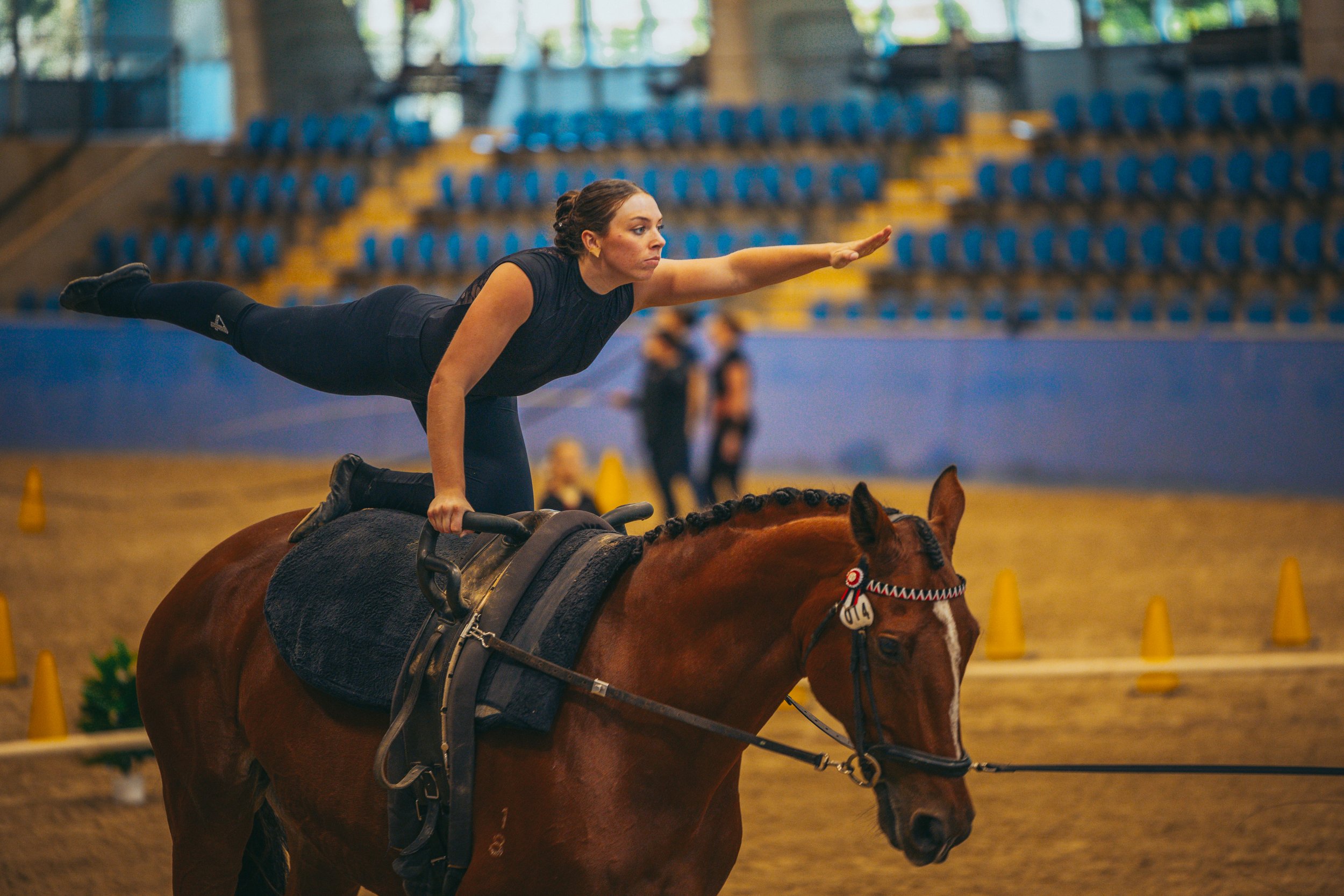 A woman practicing balancing on a galloping horse inside an indoor arena.