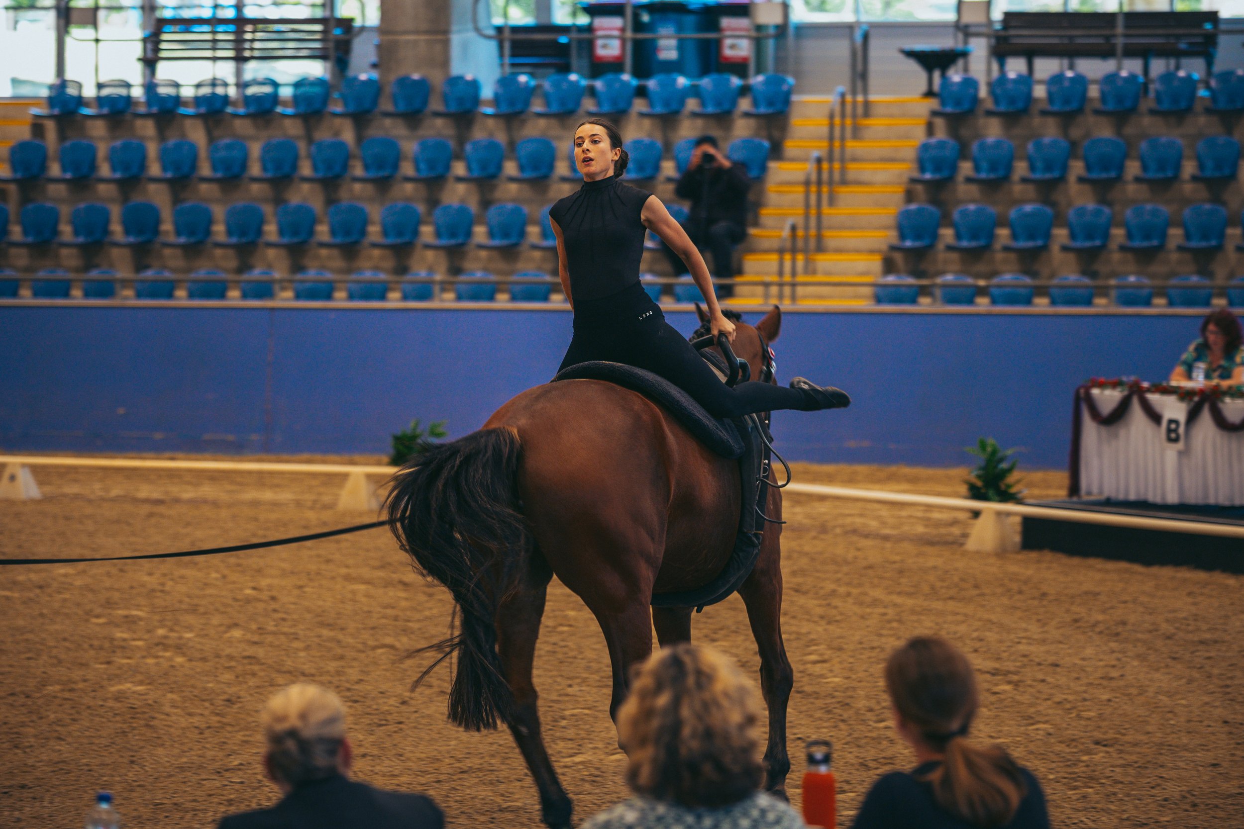 A woman performing an equestrian routine on a horse in an indoor riding arena, with spectators watching.