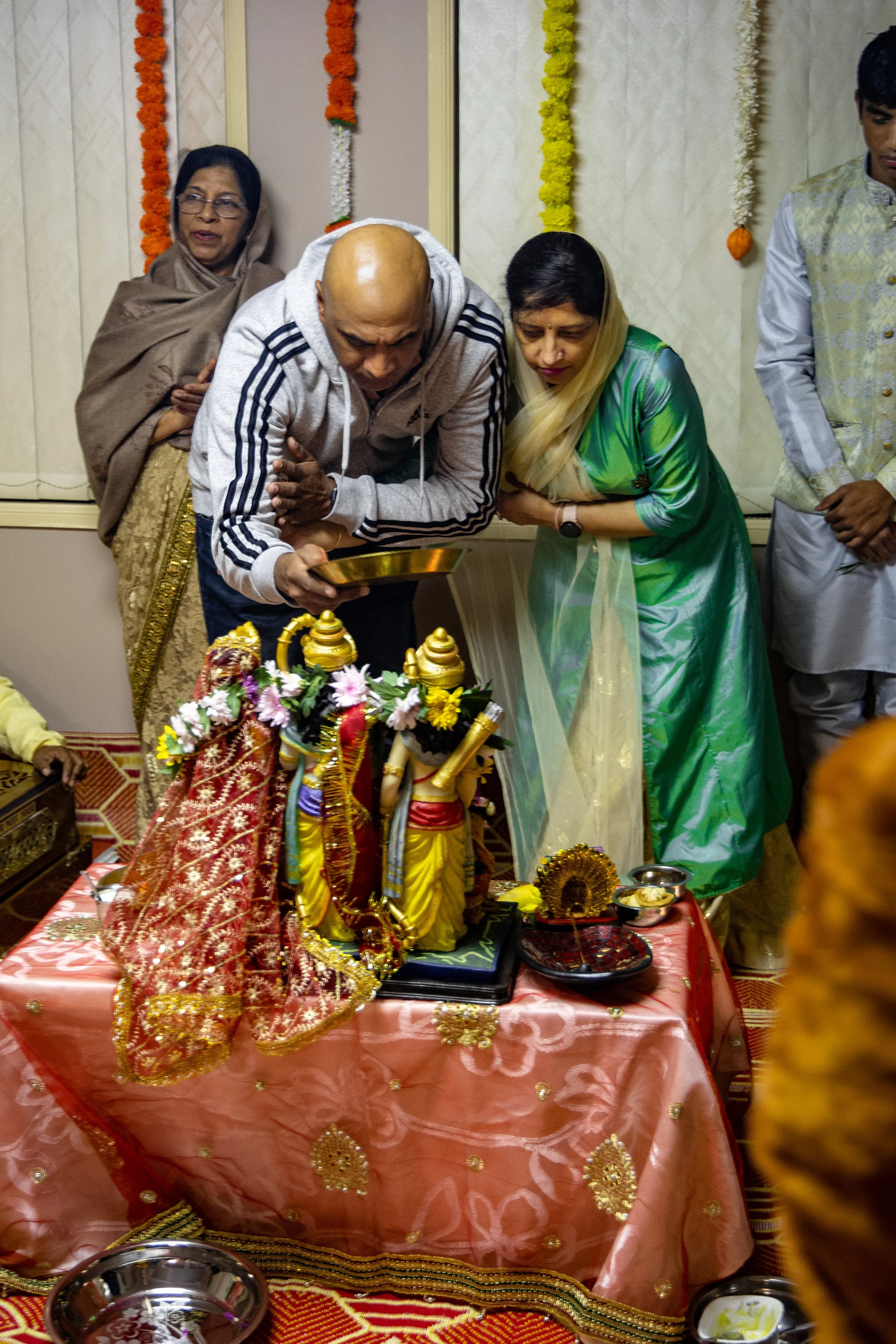 People participating in a Hindu religious prayer or ceremony, bowing before decorated statues of deities on a table covered with a pink cloth, with traditional offerings and decorations.