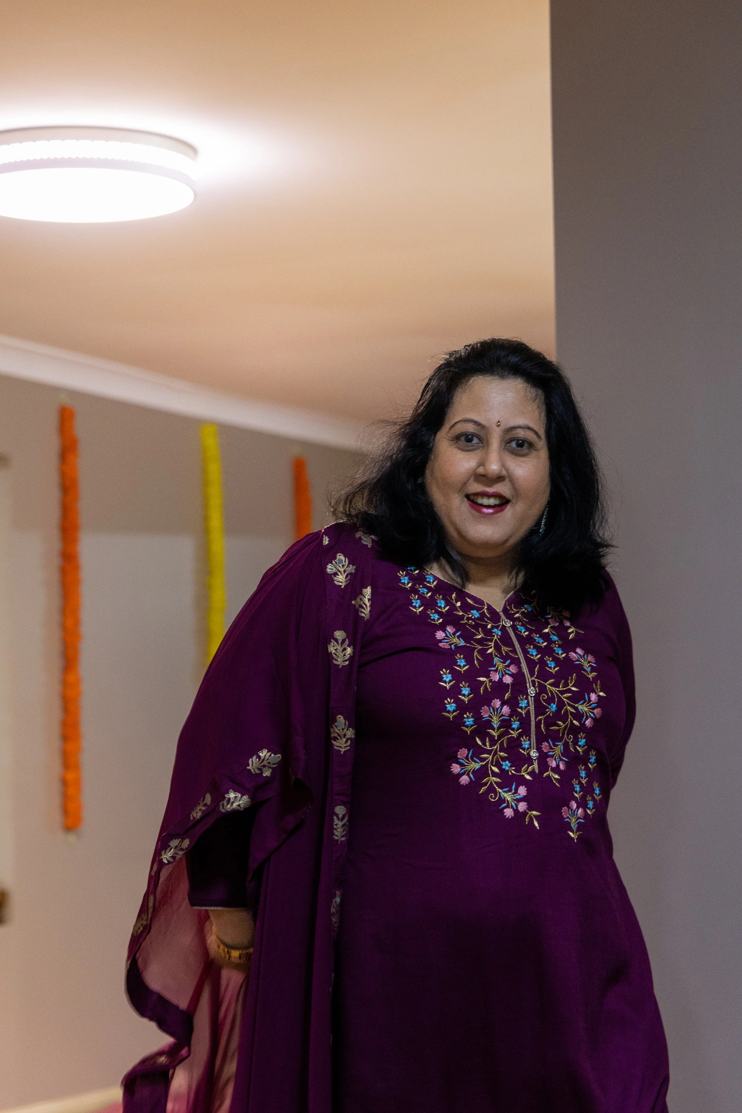A woman with dark hair wearing a purple embroidered traditional Indian dress, smiling indoors with festive decorations in the background.