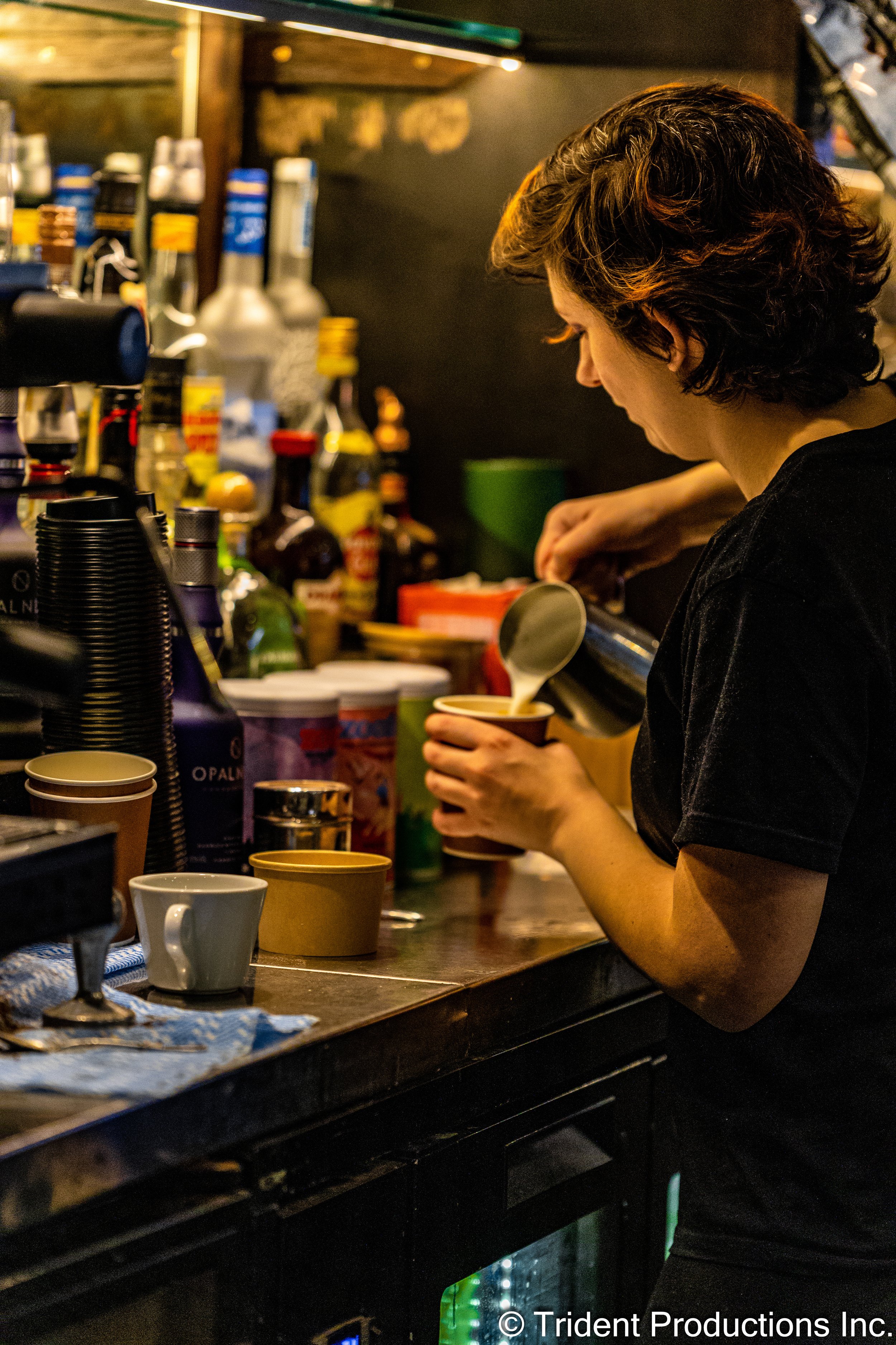 A woman with short, curly brown hair prepares a coffee drink behind a bar in a coffee shop. She is pouring steamed milk into a paper cup.