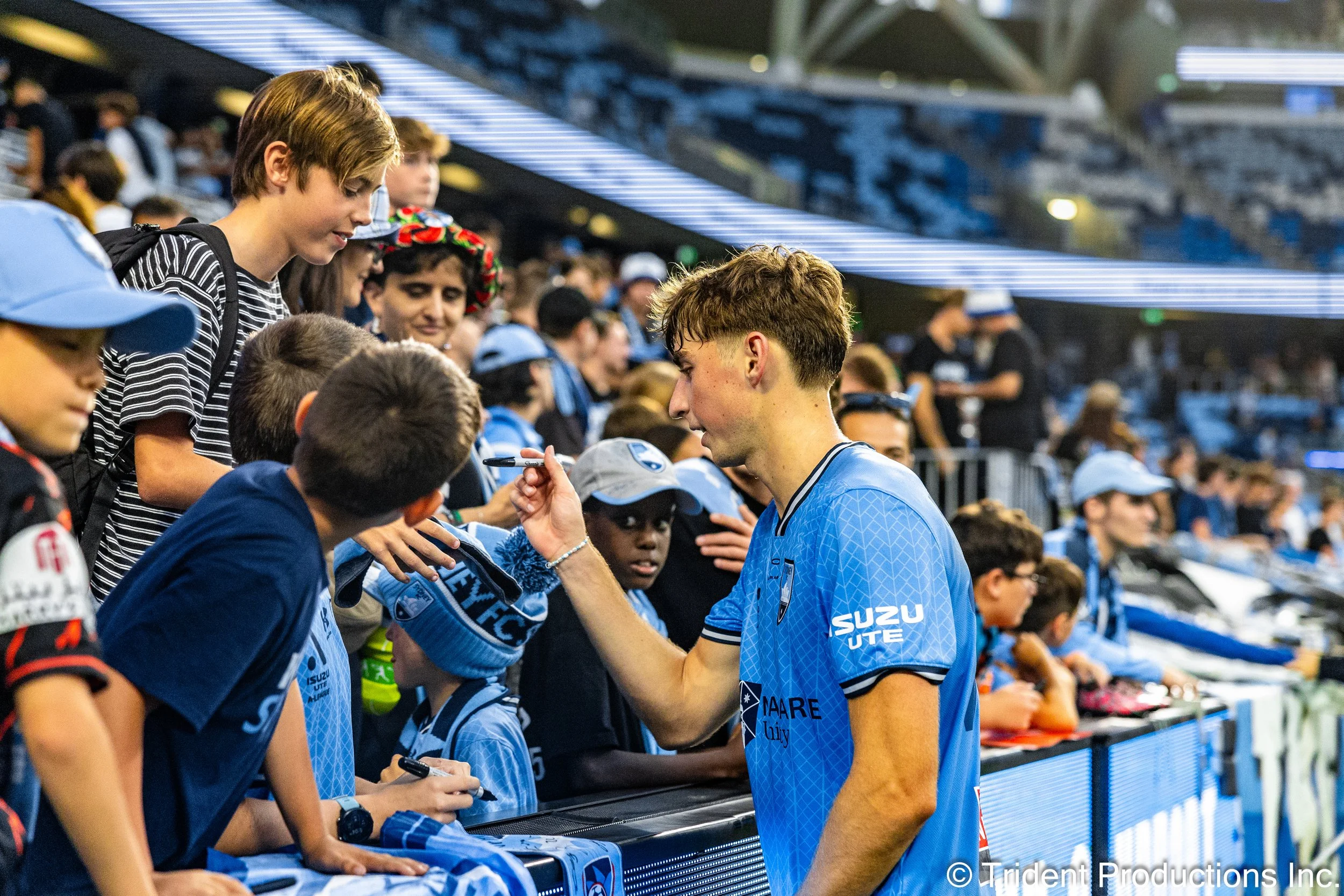 A young male athlete in a blue sports jersey signing autographs for children and fans at a stadium.
