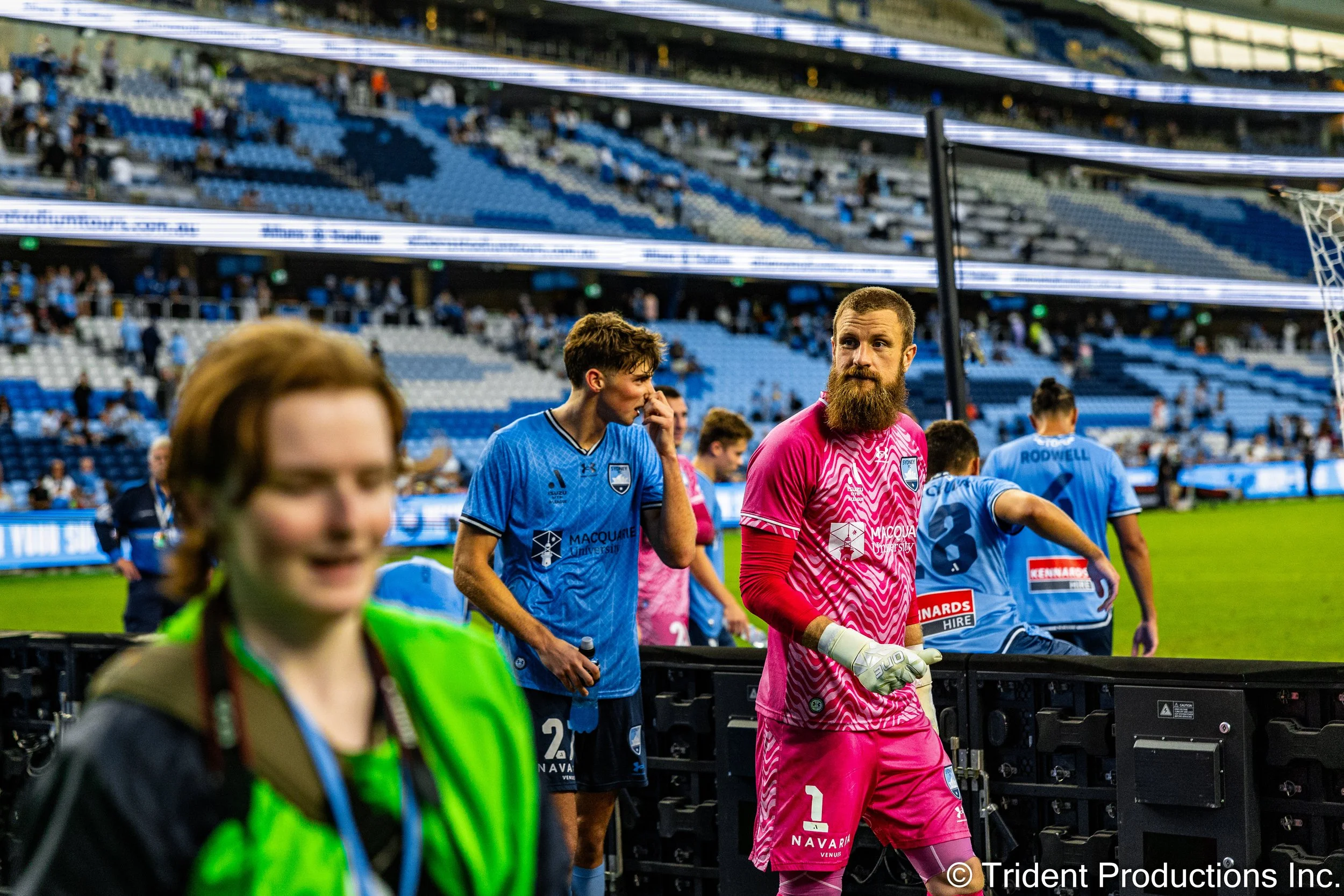 Soccer players on field after match, one in pink goalkeeper uniform and others in blue jerseys, with stadium audience in background.