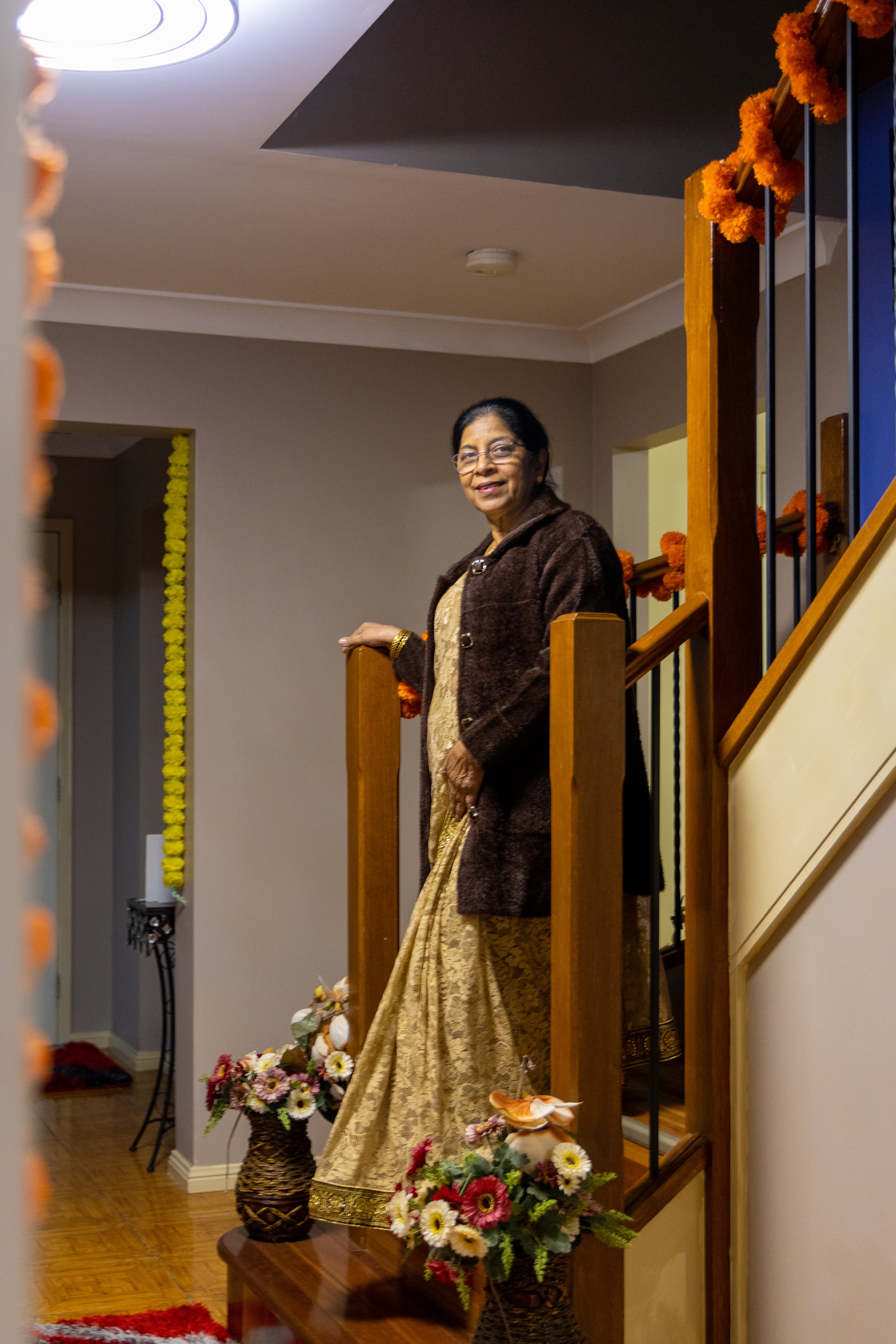 An elderly woman standing on a staircase, smiling, dressed in traditional Indian attire with a brown coat. The area is decorated with orange marigold flowers, and flower arrangements are at the base of the stairs.
