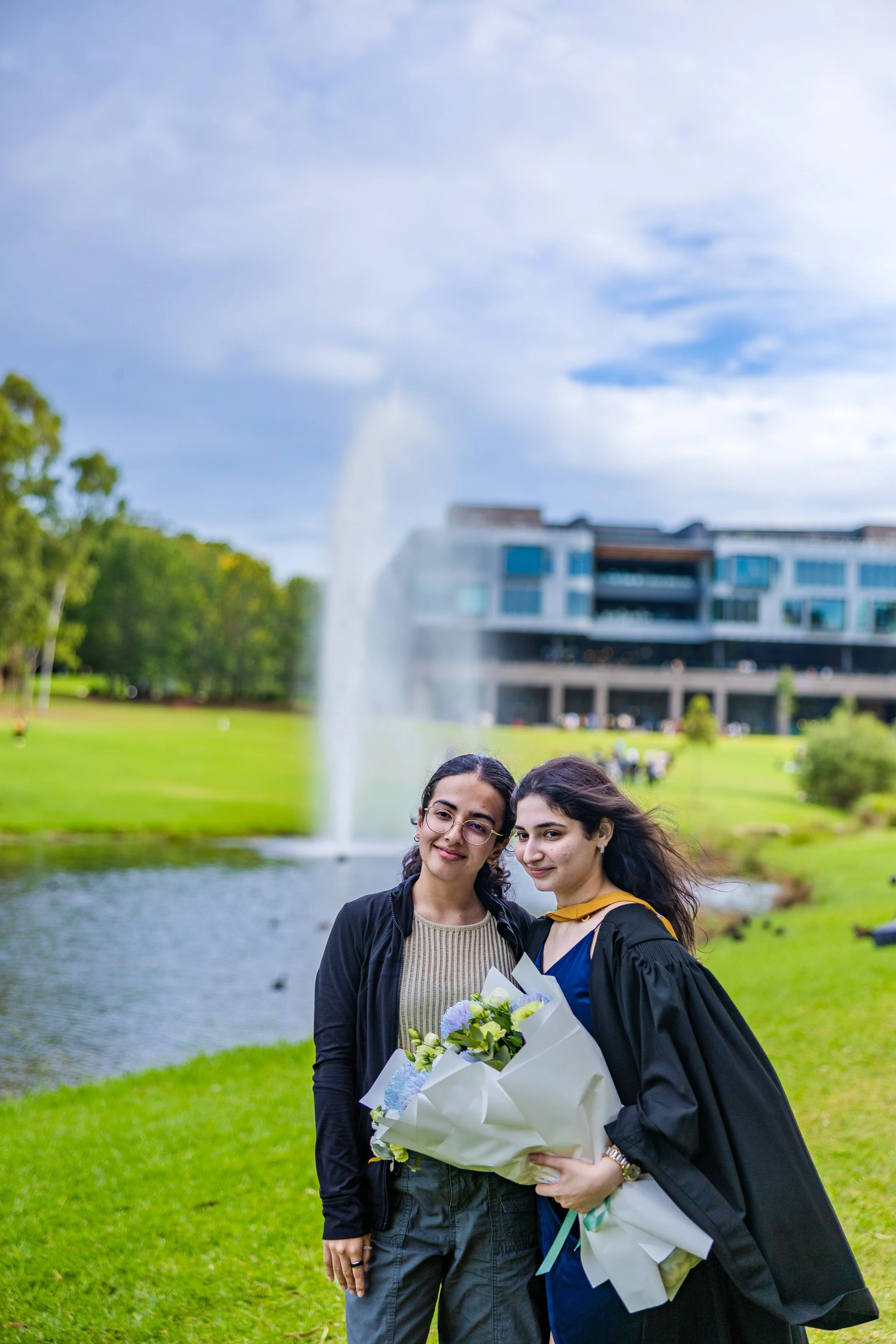Two women standing outdoors by a pond, one in graduation attire and holding a bouquet of flowers, smiling at the camera.