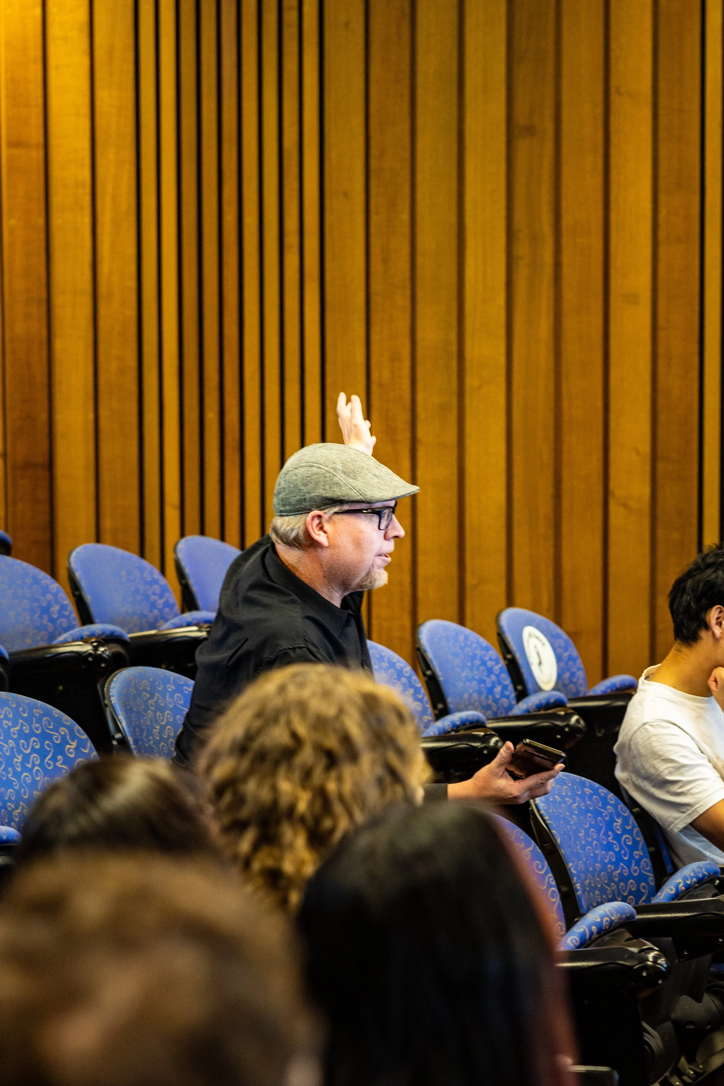 A man wearing a gray flat cap, glasses, and black shirt sitting in a blue auditorium seat, holding a smartphone, with an empty seat in front of him.
