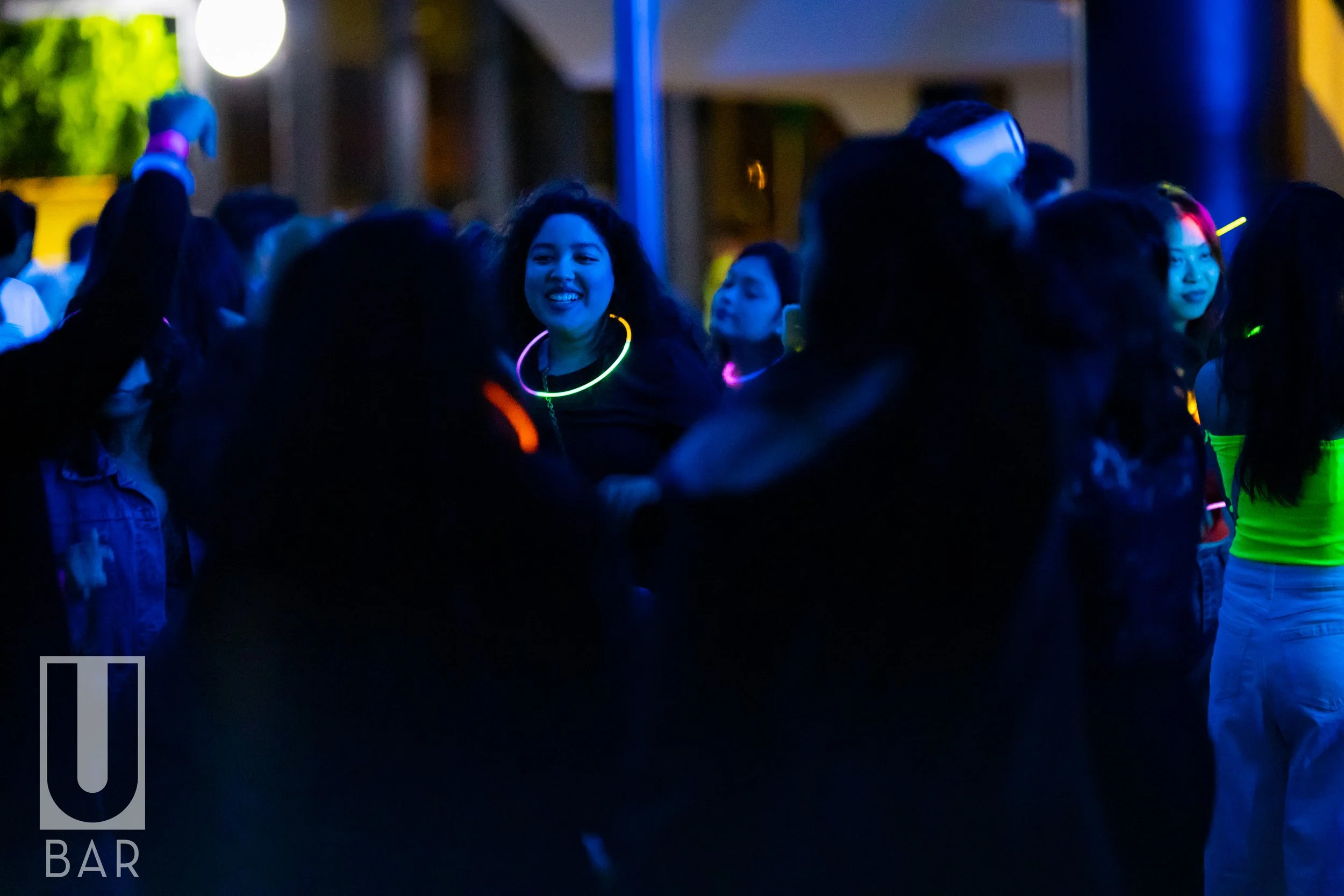 People dancing at a party with glow-in-the-dark accessories in a dimly lit venue, with a woman smiling and wearing a glow necklace.