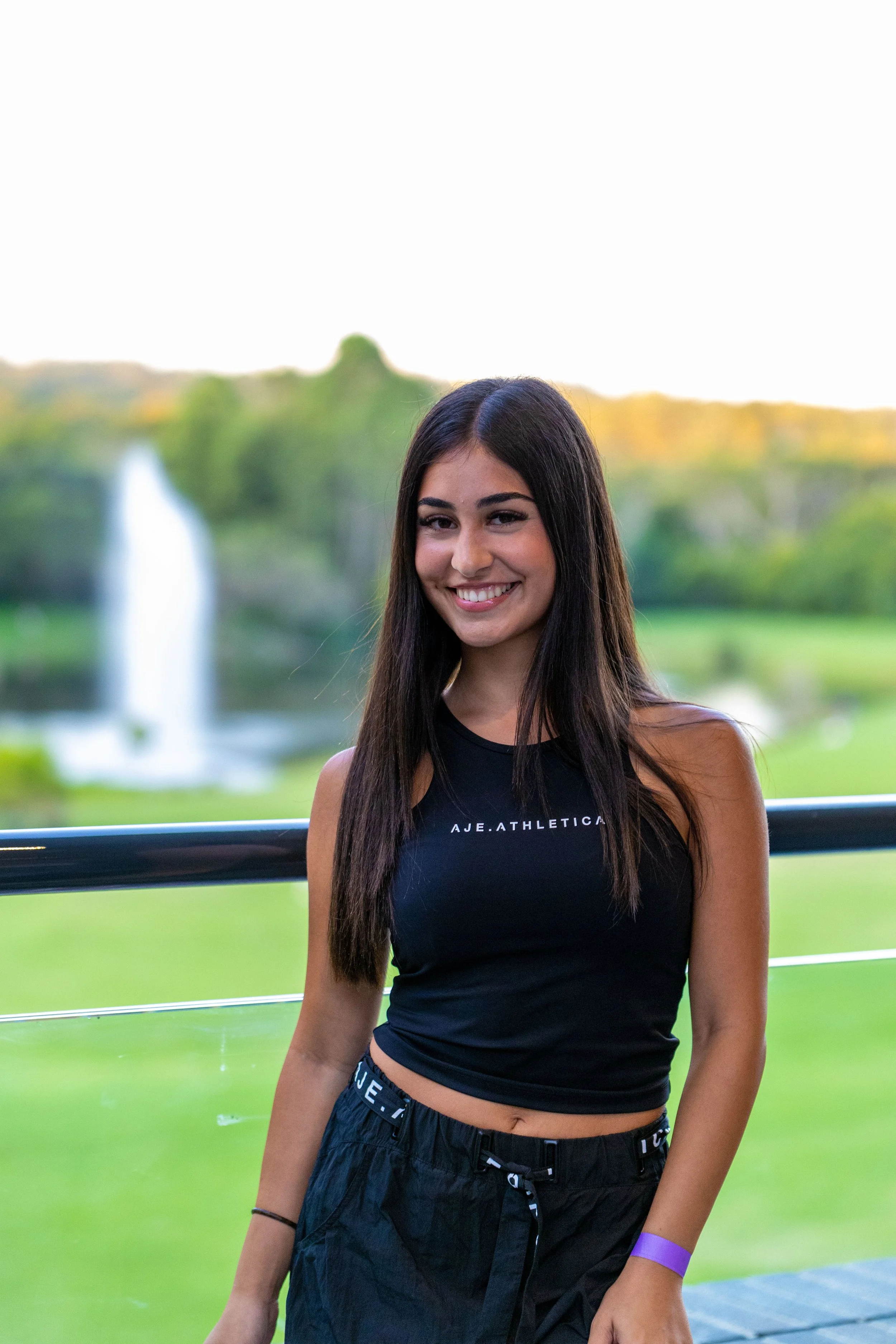 A young woman with long dark hair smiling outdoors in front of a park with a fountain and trees.