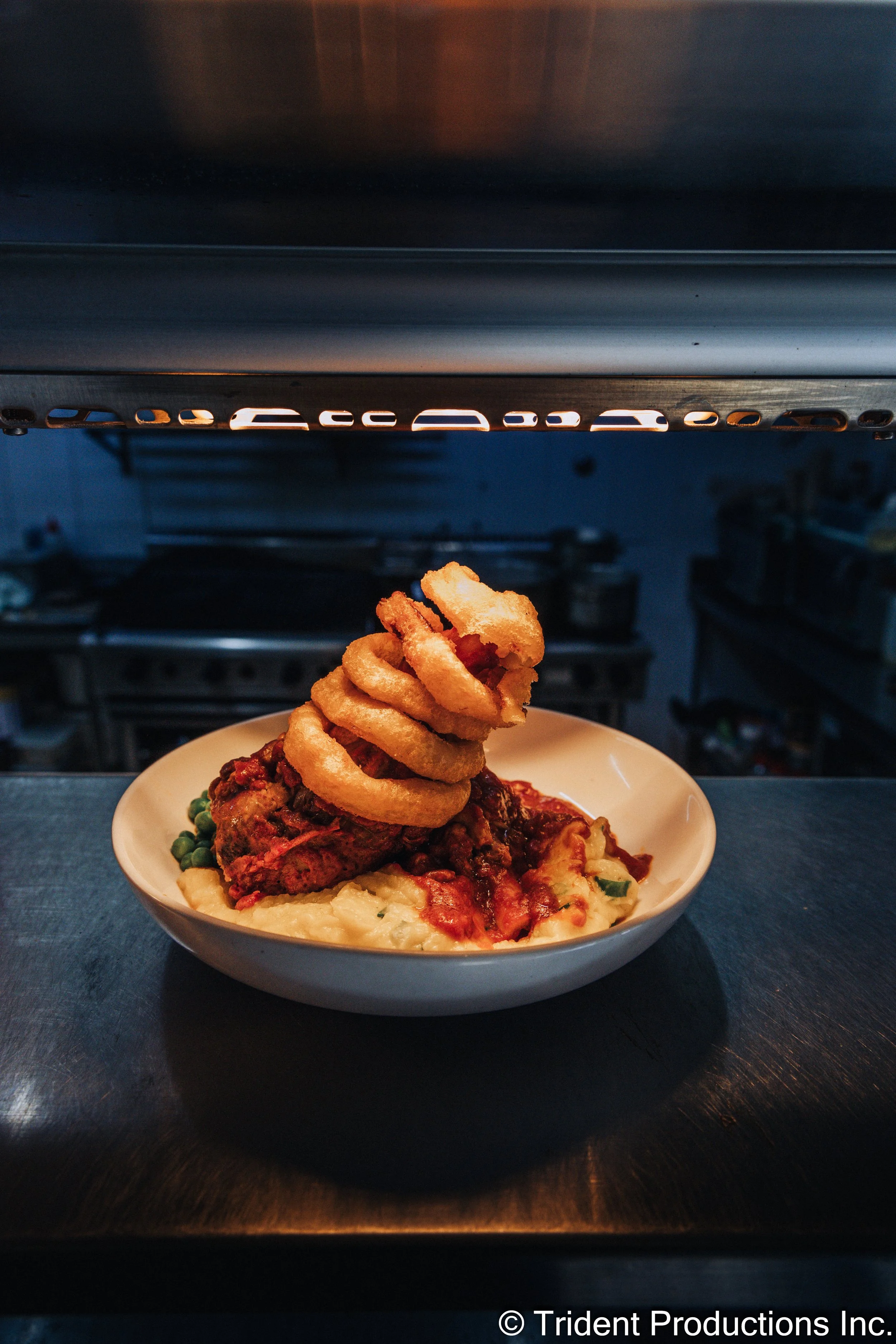 A bowl of mashed potatoes topped with meatloaf, onion rings, and green peas, placed on a dark table with a kitchen in the background.