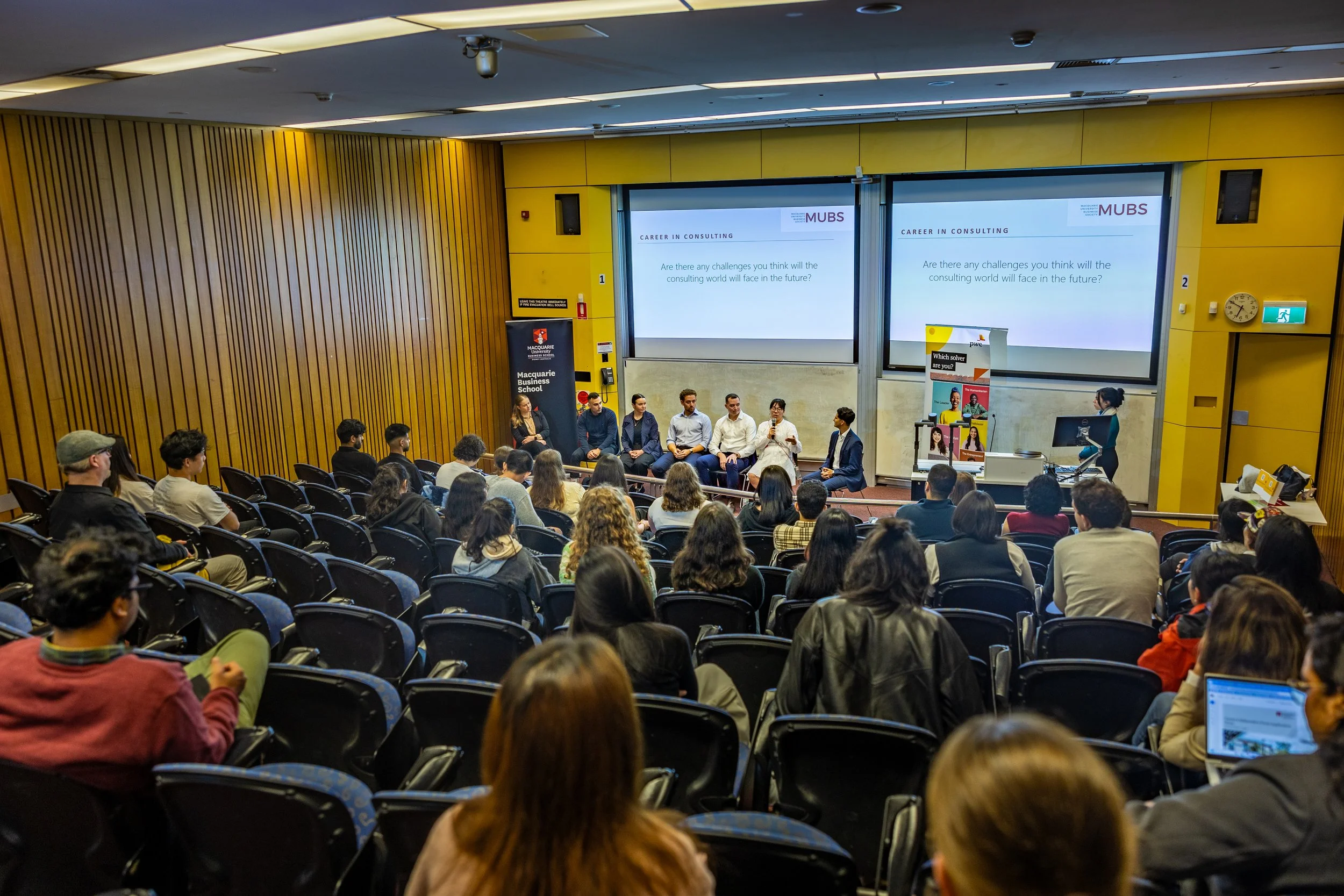A panel of seven speakers seated on stage in a lecture hall, with a woman standing and speaking to an audience seated in black chairs. The screens behind display a presentation titled 'Career in Consulting' from MUBS. The hall has wooden paneling on 