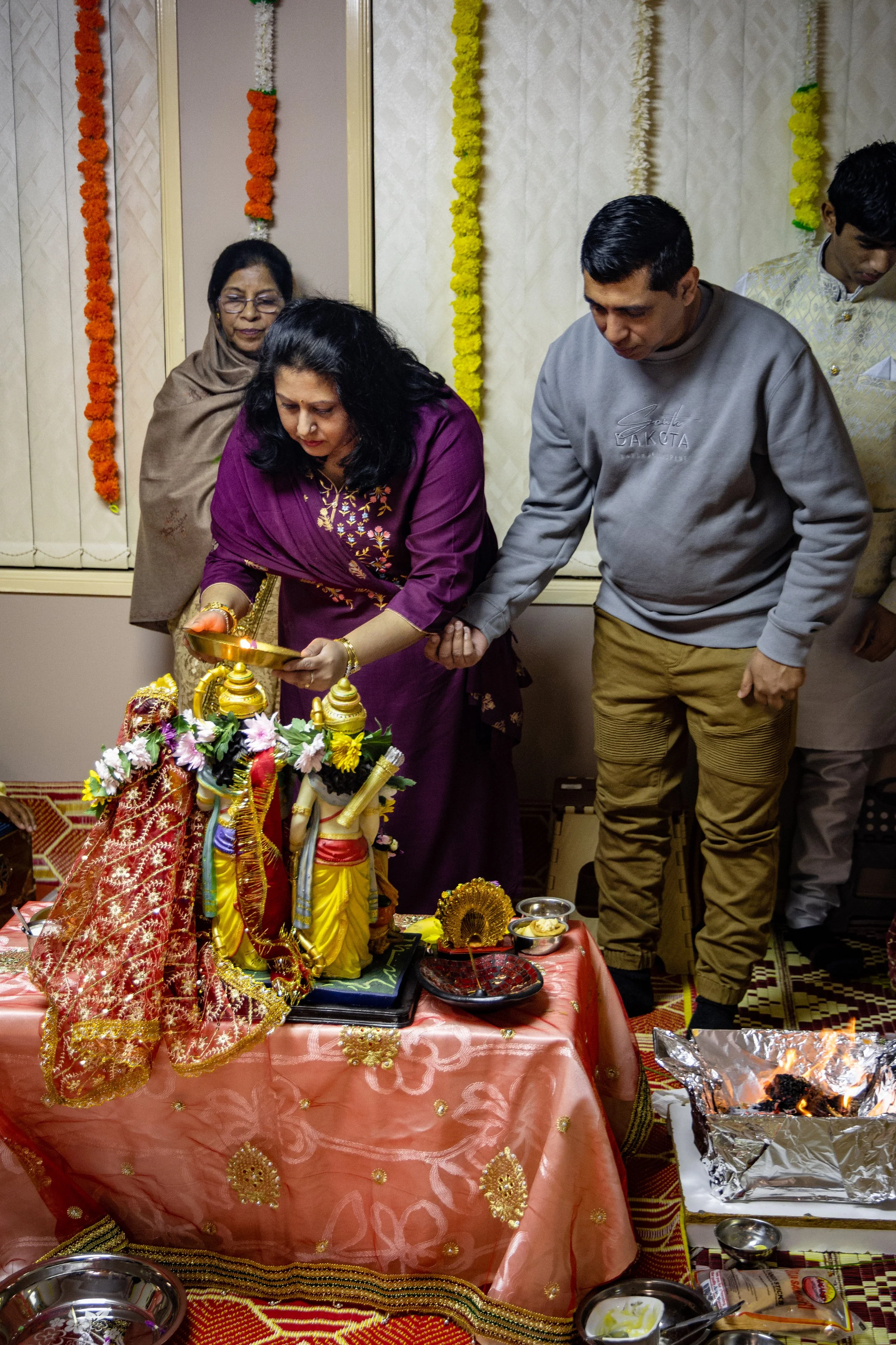 People perform a Hindu prayer ritual at a decorated altar with statues of deities, flower garlands, and offerings.