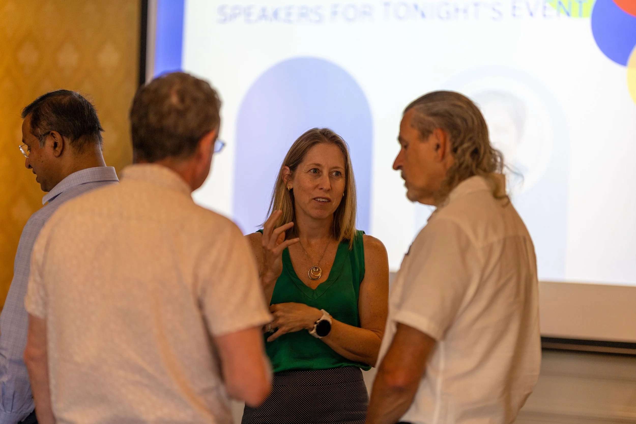 Four people in a conversation at an event, with a presentation screen in the background displaying the text 'Speakers for Tonight's Event'