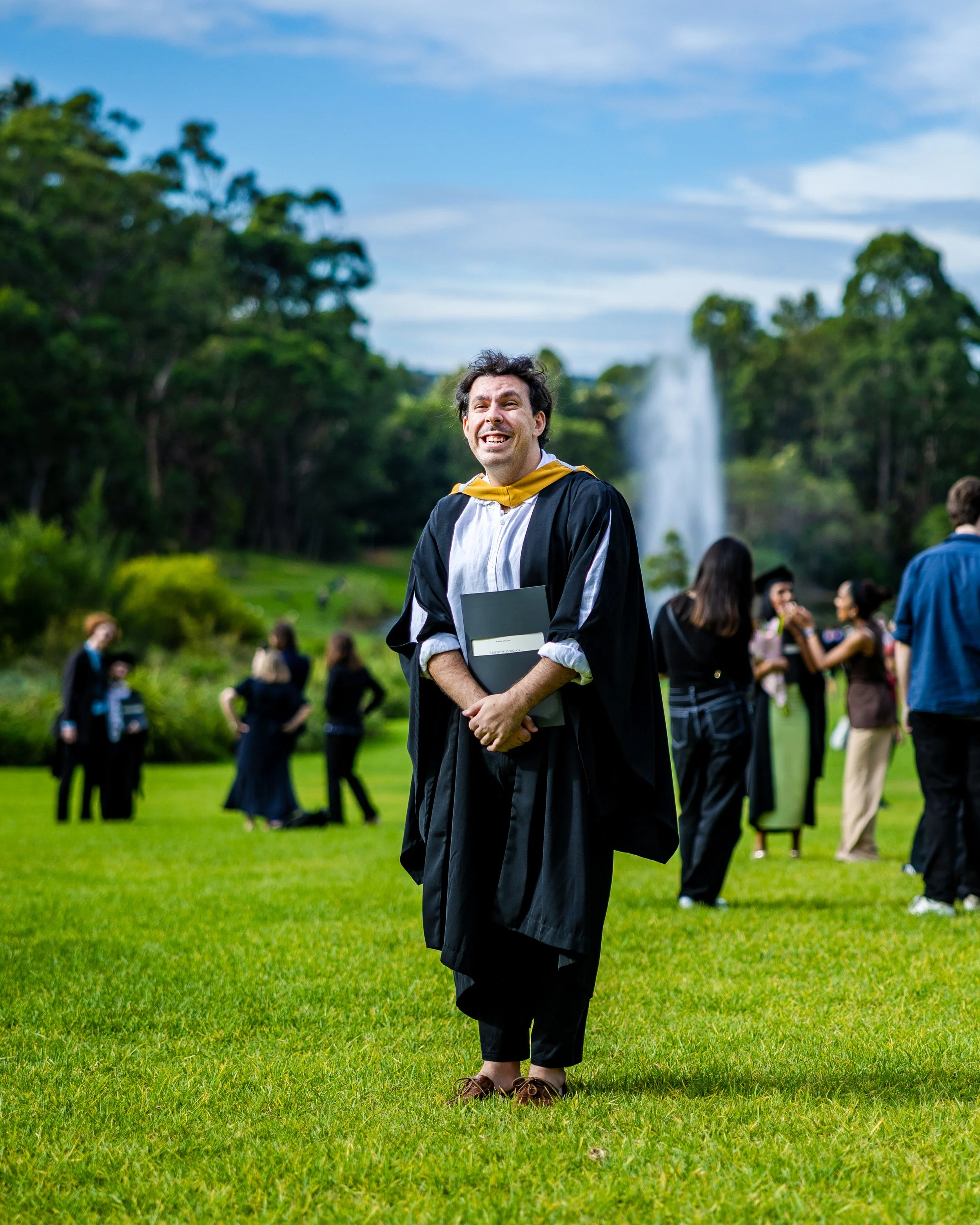 A man in a graduation gown and cap holding a diploma, smiling outdoors at a graduation ceremony with other graduates and a fountain in the background.