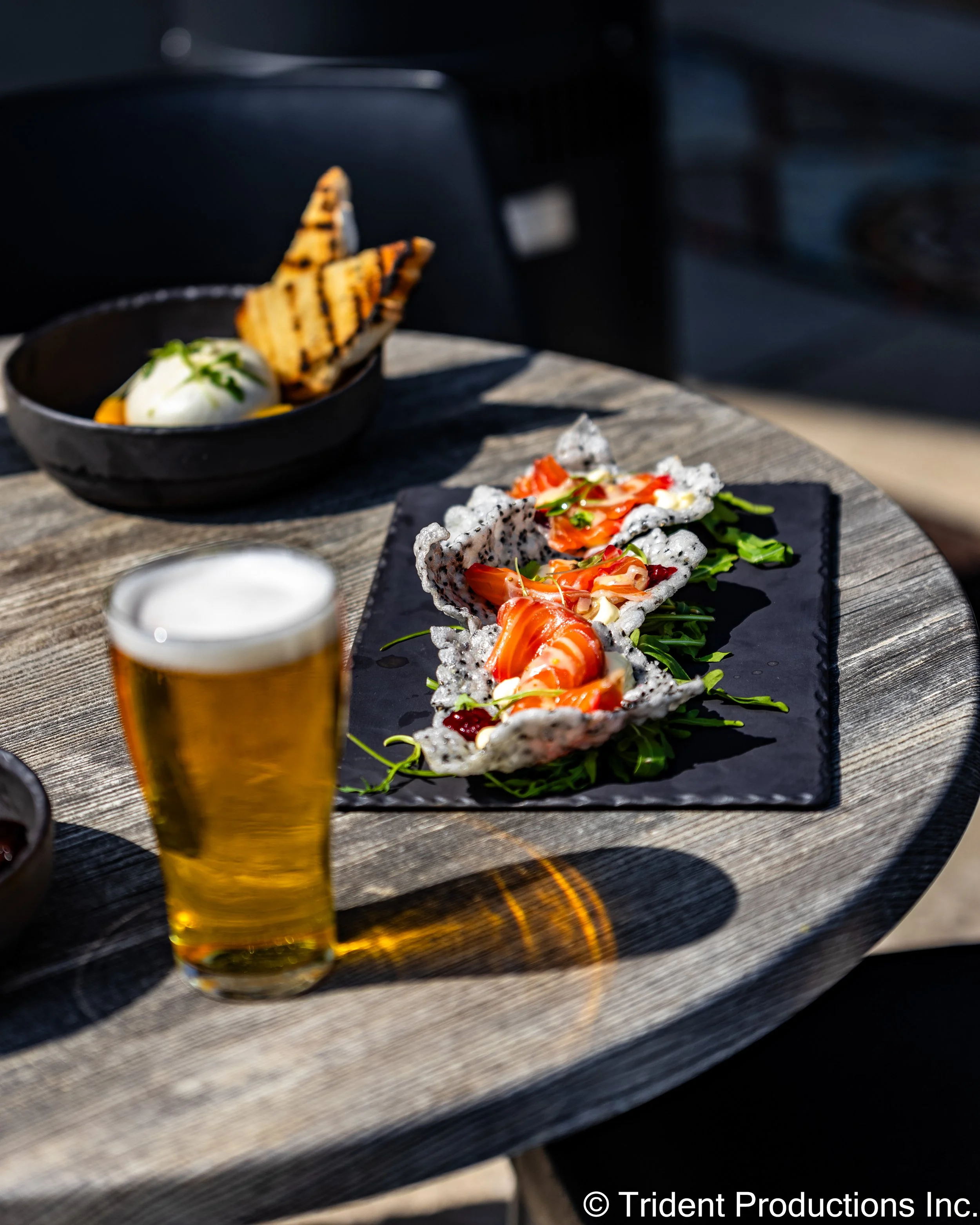 A glass of beer, a dish with smoked salmon, and a bowl with grilled bread and a poached egg on a wooden outdoor table.