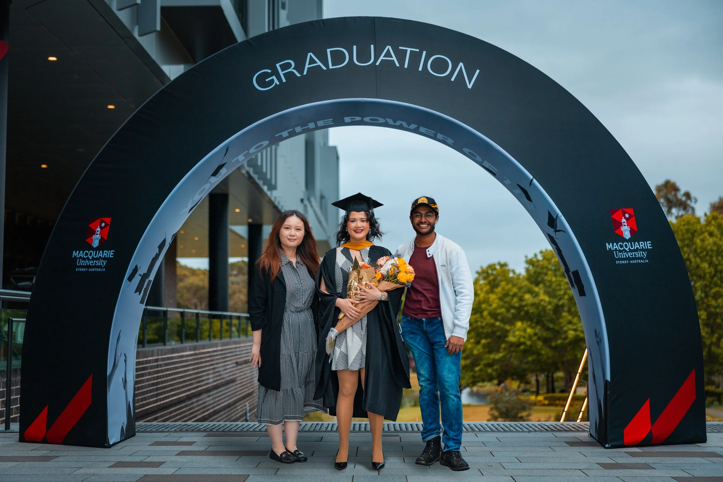 A joyful woman in graduation cap and gown holding a bouquet of flowers, standing with two friends under a large archway with 'Graduation' and 'The Power of Education' written on it, outside at Macquarie University in Sydney, Australia during daytime.
