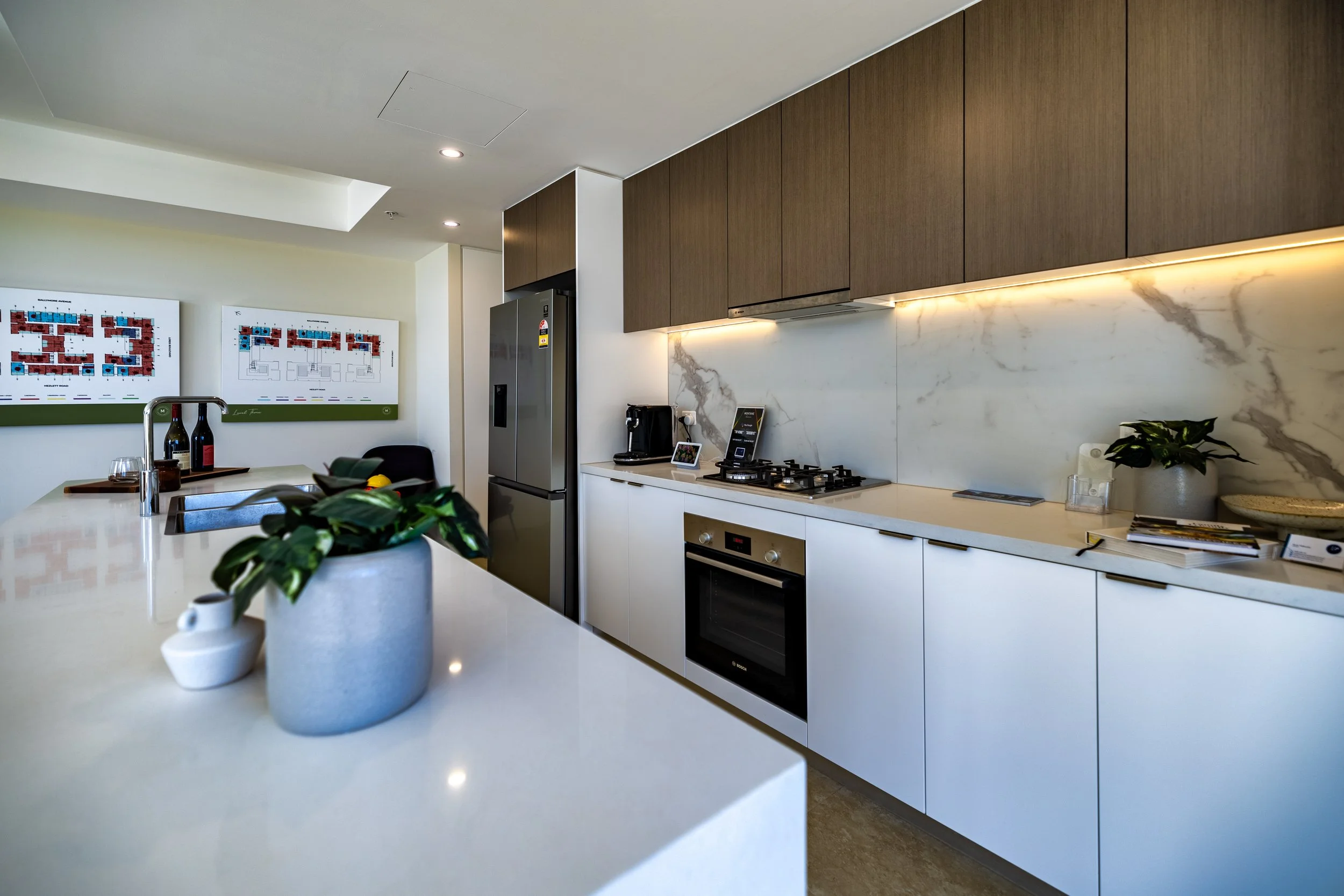 Modern kitchen with white countertops, wooden upper cabinets, marble backsplash, stainless steel refrigerator, oven, and stove; potted plants and kitchen accessories on the counters; dining area with framed artwork on the wall.