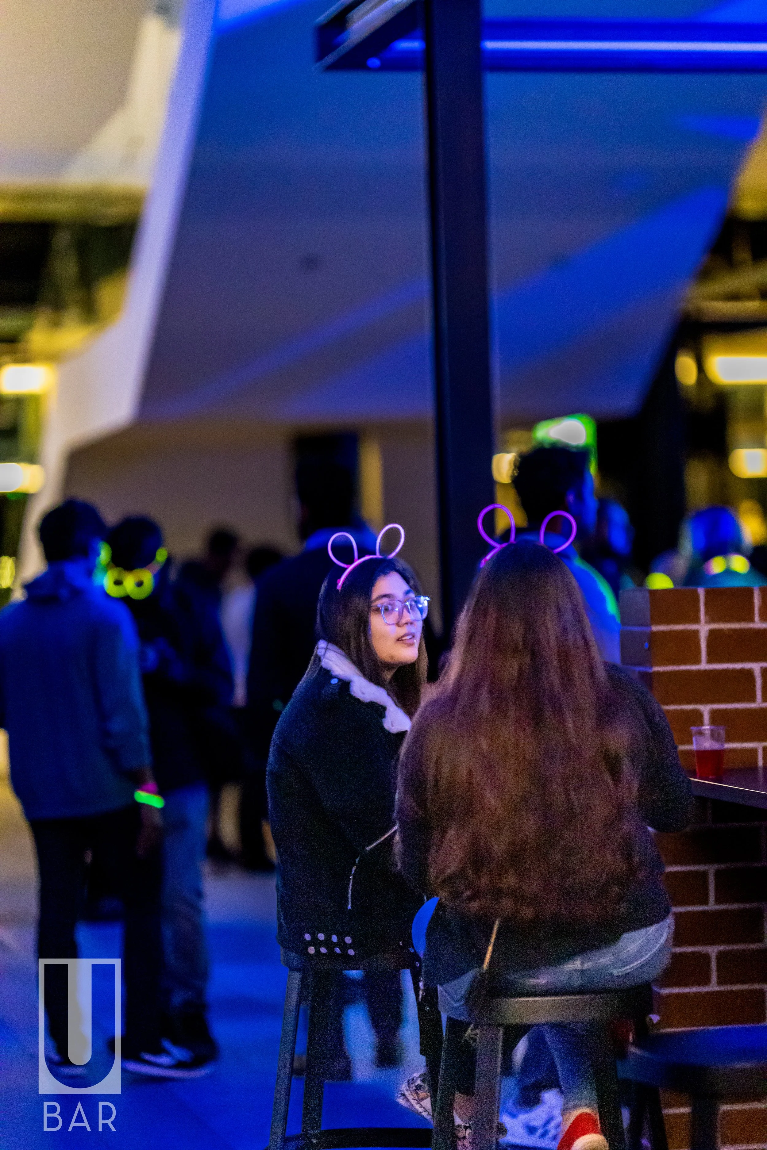 Two women with glowing deer antlers headbands sitting at a bar, talking, with people dancing in the background at night.