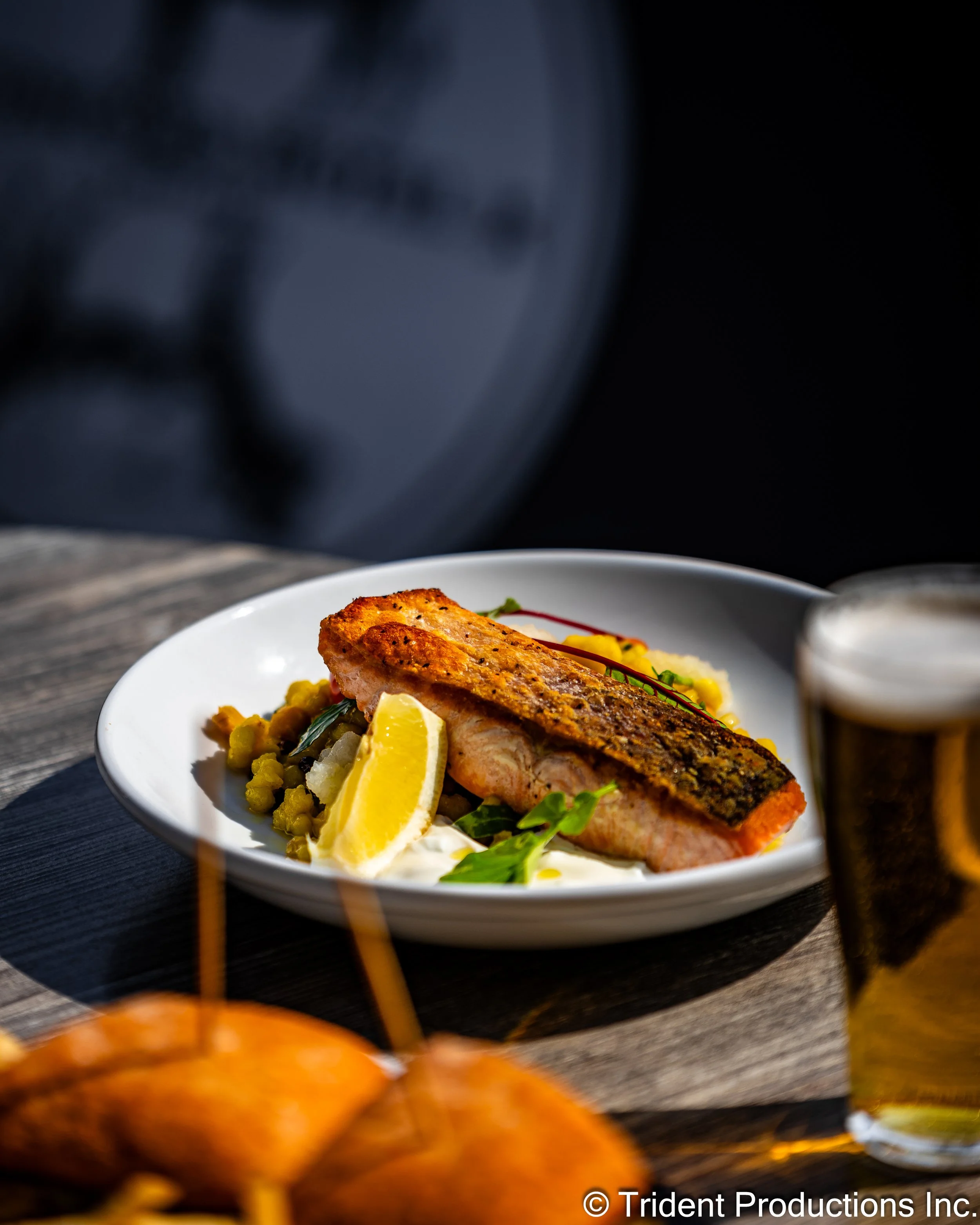 A plate with grilled salmon, a lemon wedge, and garnishes on a wooden table, with a glass of beer in the background.