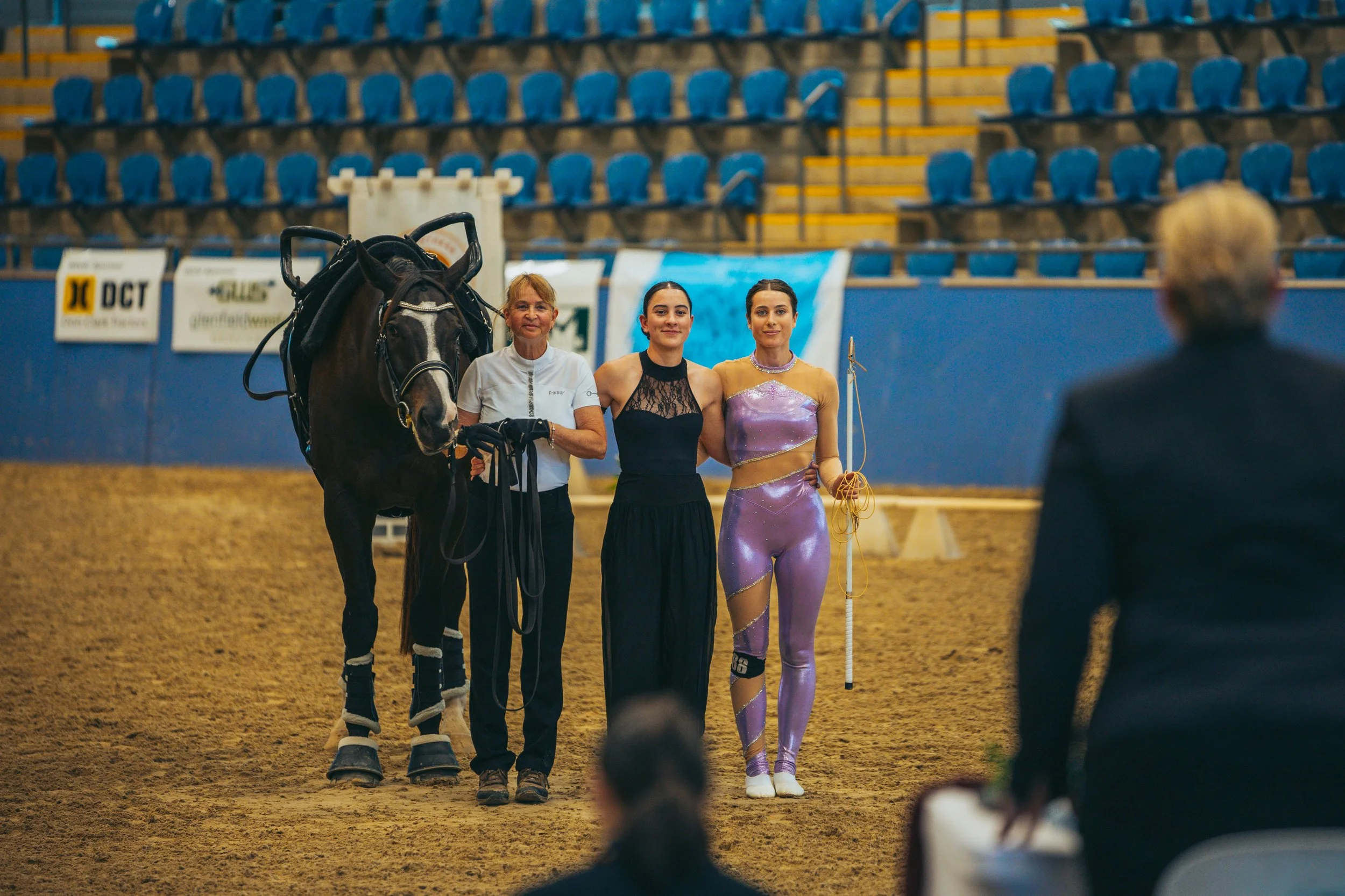 Three women and a horse standing in an indoor arena, with an audience and blue stadium seats in the background. One woman is dressed in athletic wear, another in a black dress, and the third in a shiny purple rhythmic gymnastics costume holding a clu