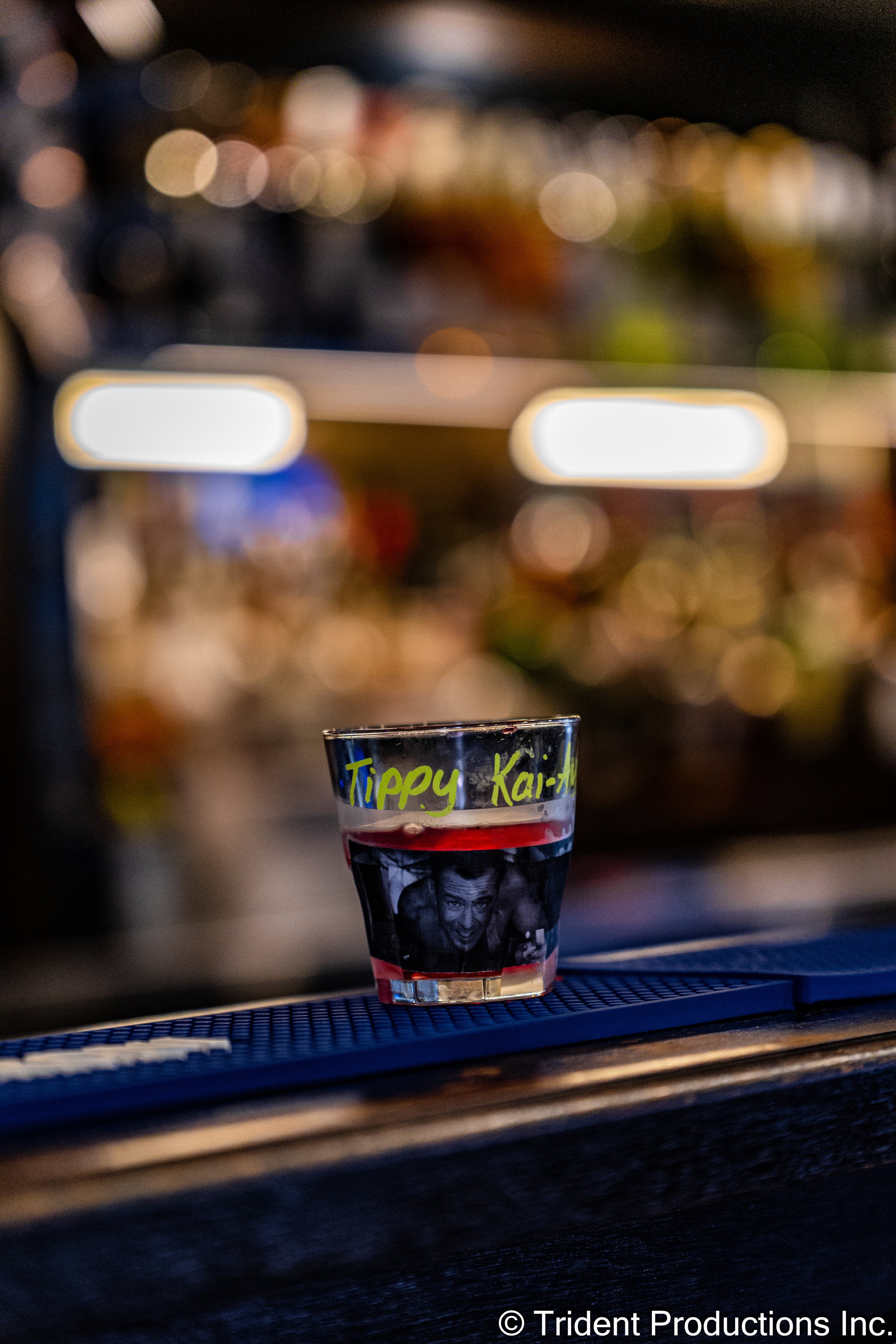A shot of a glass filled with a red drink, sitting on a bar counter, with the words "Tippy Kai" written on it in neon yellow marker, and a photo of a man's face underneath, with a blurry background of bar shelves.