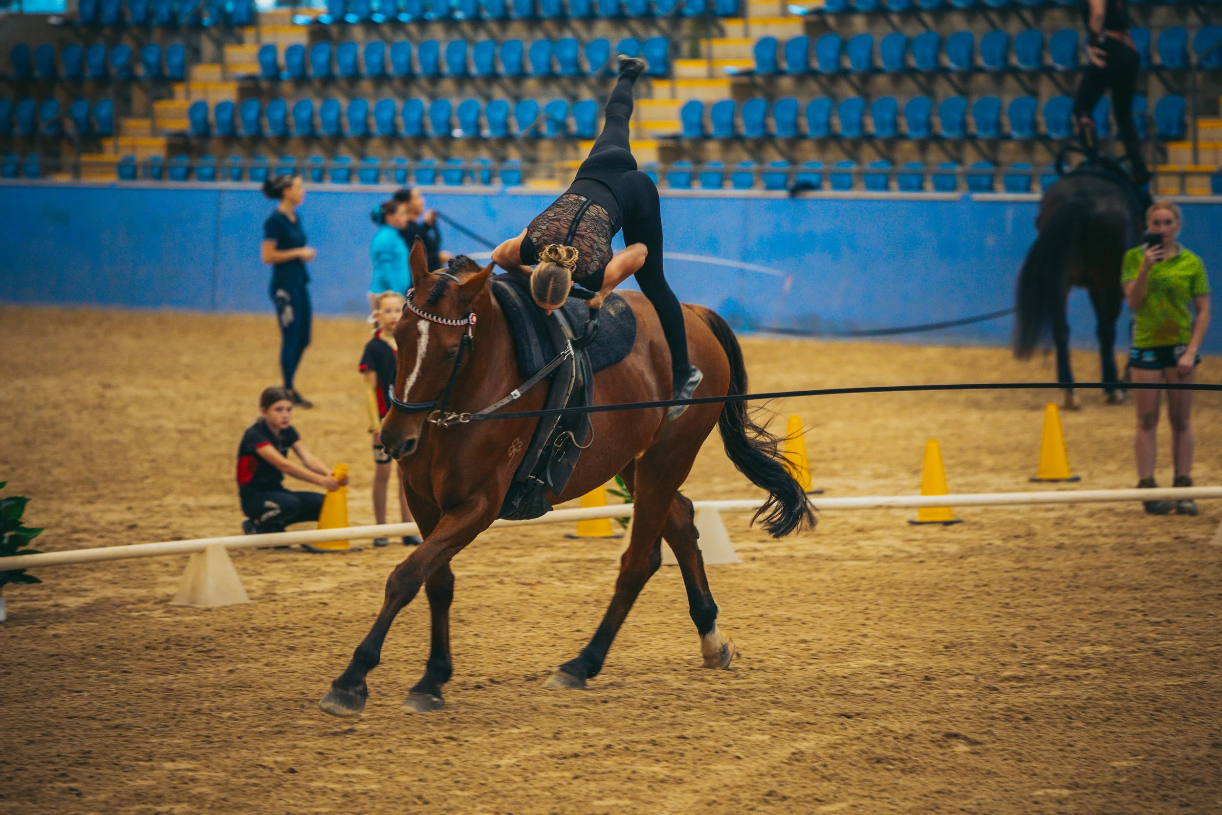 A woman in black performing a ballet dance on horseback inside an indoor arena, with people and other horses in the background.
