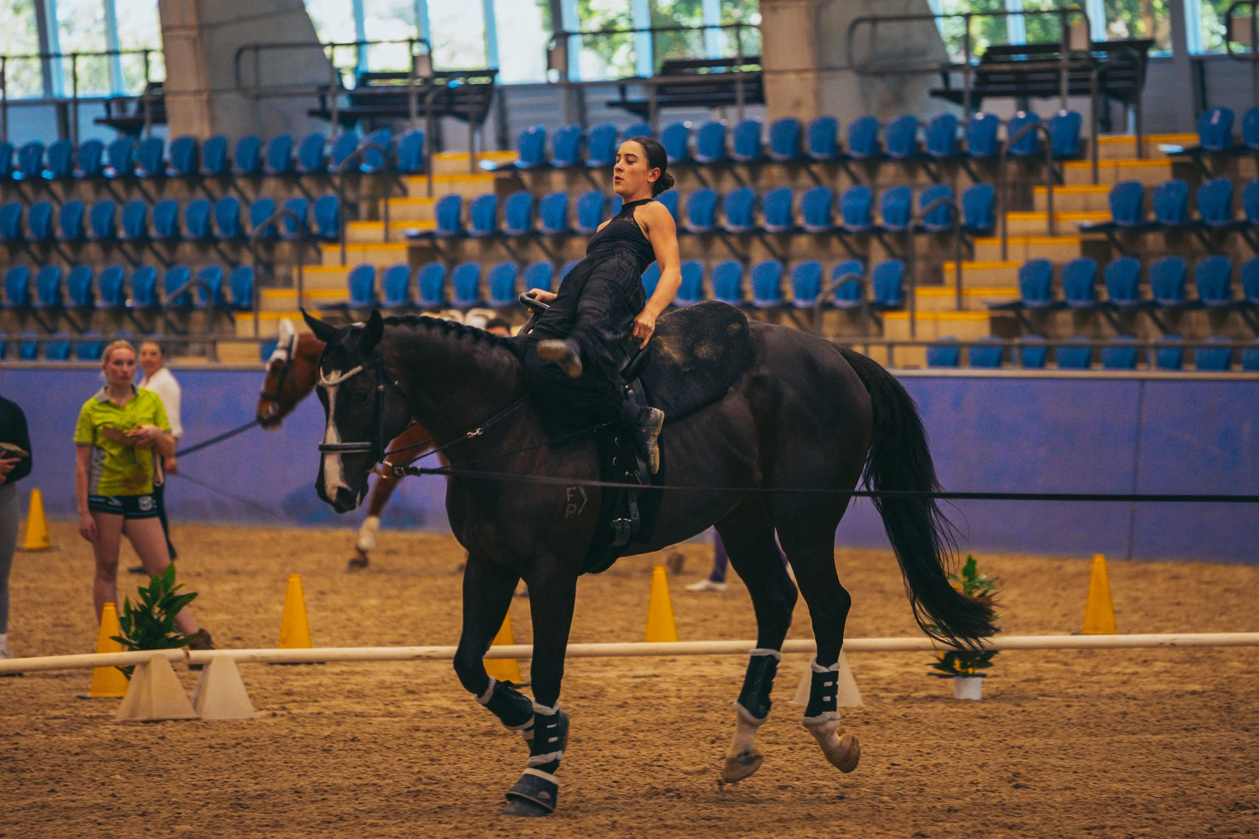 A woman in black riding attire riding a dark horse in an indoor arena, with other riders and spectators in the background.